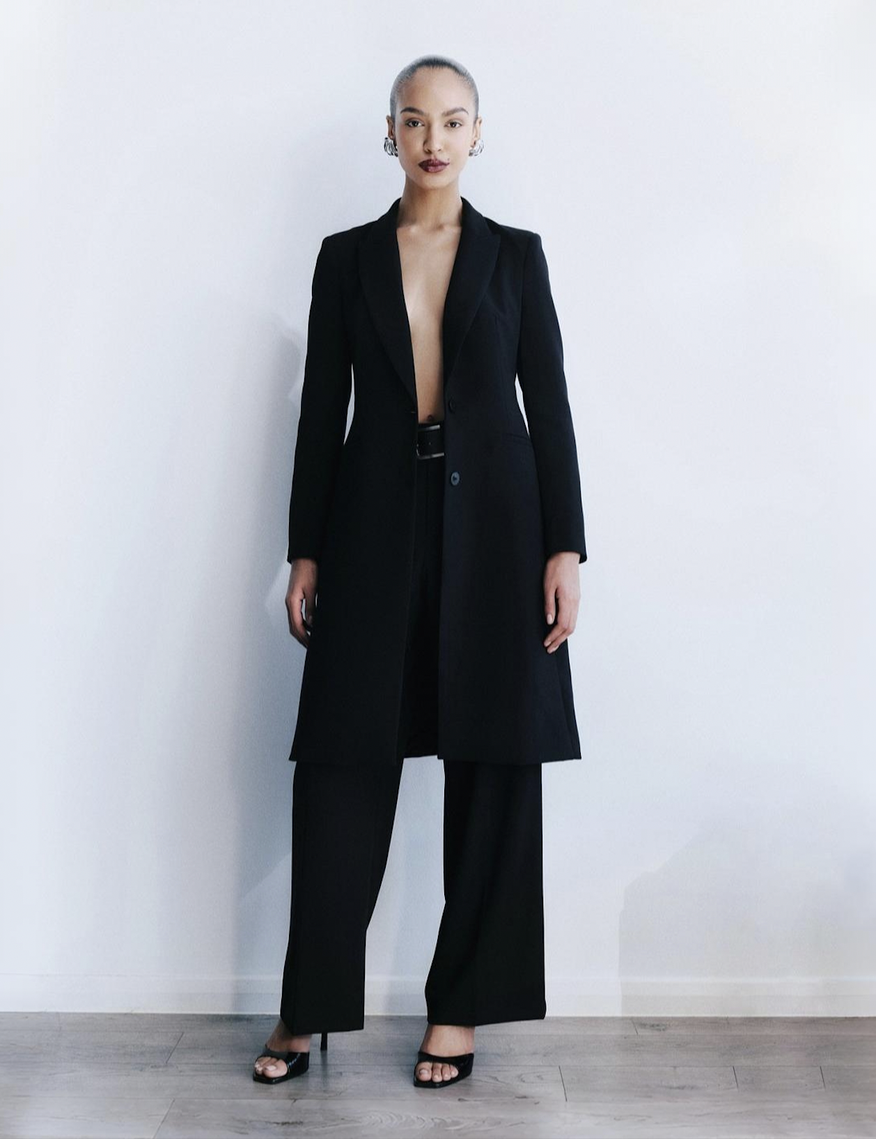 A model with a slick bun hairstyle and dark lipstick, wearing a long black blazer with no shirt underneath, black pants, and high heels, standing against a white wall.