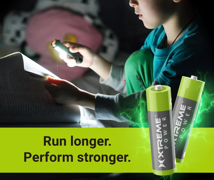 A young person using a flashlight to read a book in a dark room, with a pink pillow in the background. An overlay with two batteries and the text 'Run longer. Perform stronger.' promotes Xtreme Power batteries.