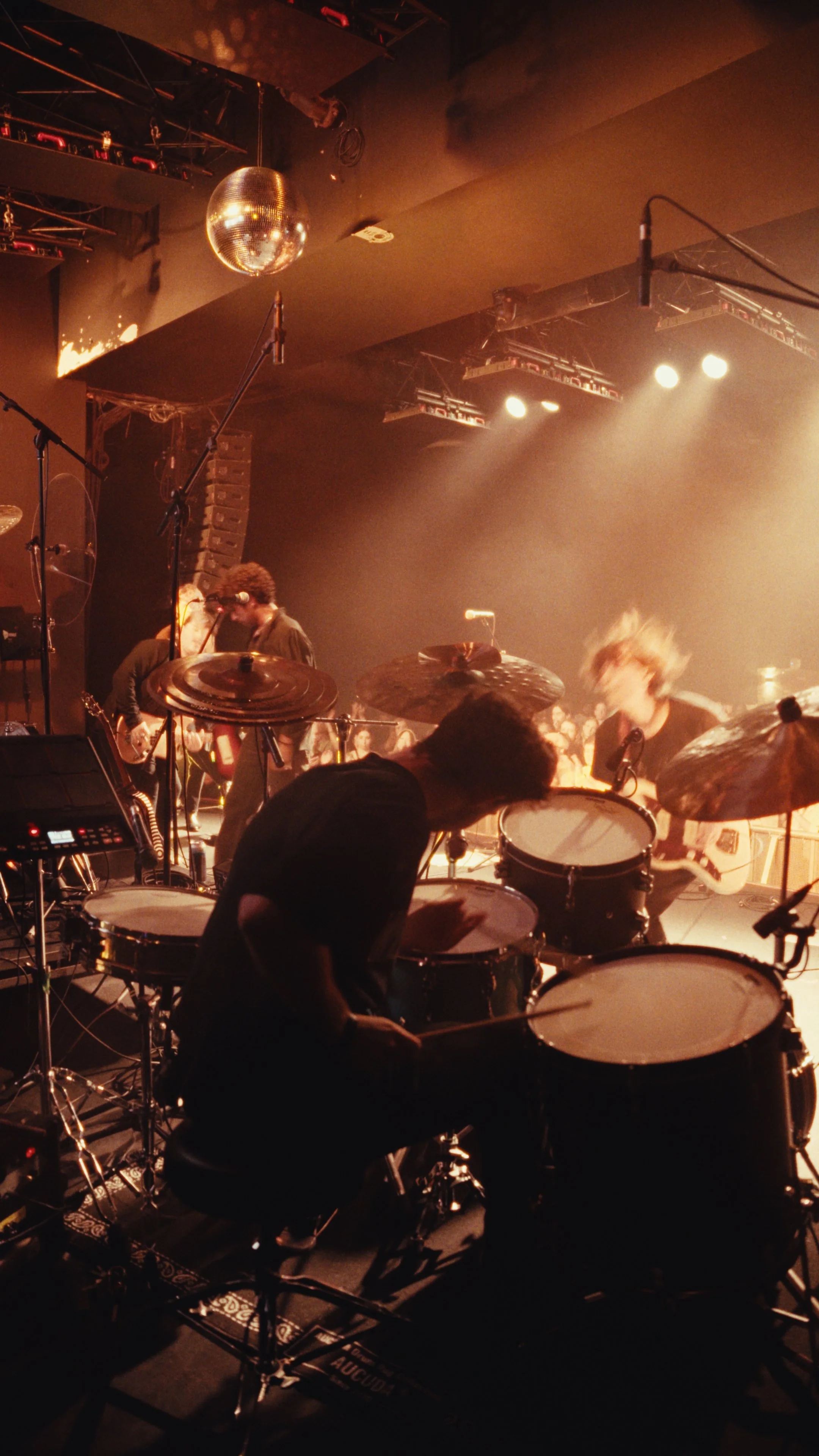 Drummer Miles Johnson performing on stage with Rohin at the Mod Club, other musicians in the background, stage lighting, and a disco ball overhead.