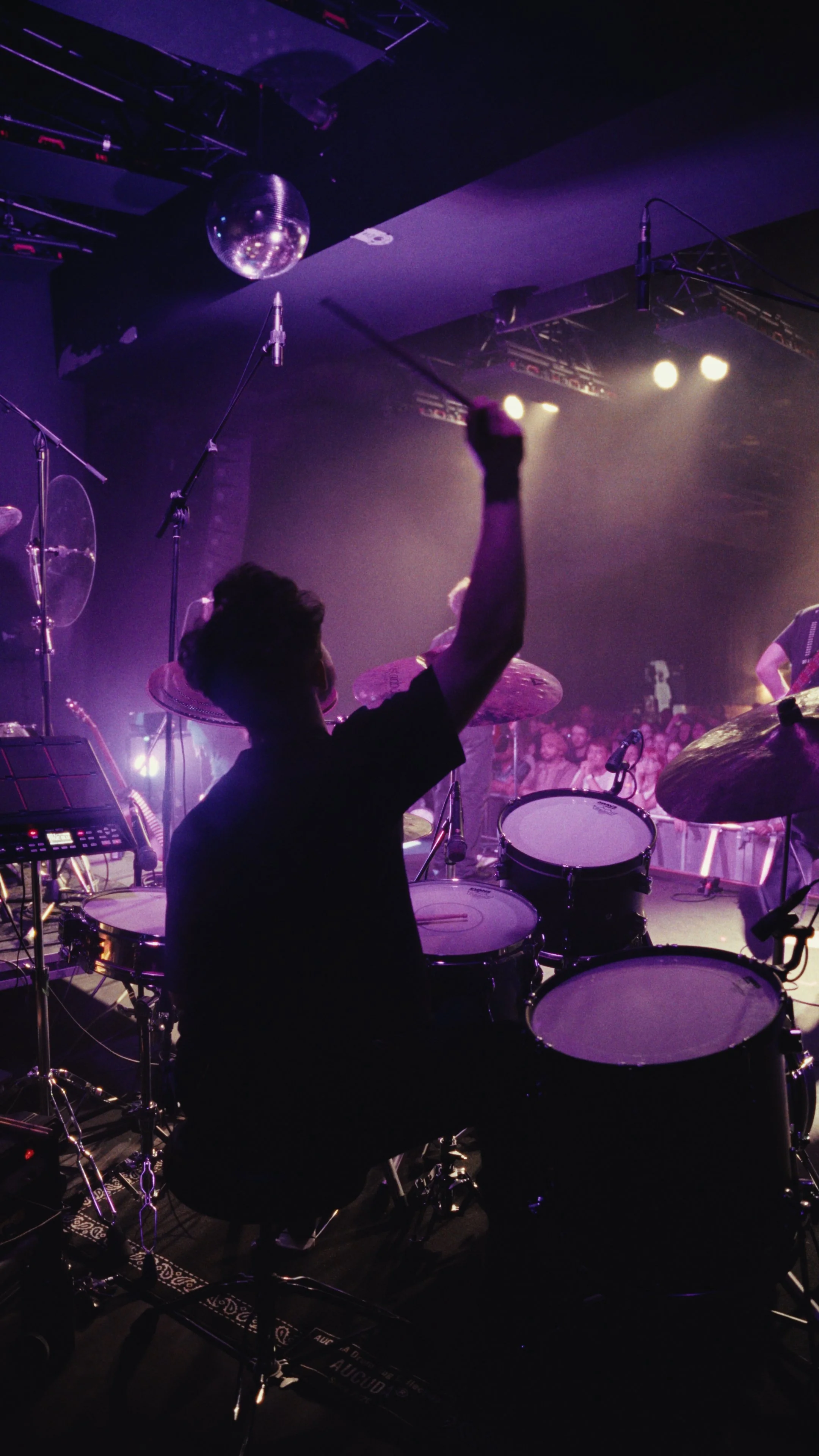 A drummer raising his fist on stage with a band performing in a dimly lit concert venue, audience visible in the background.