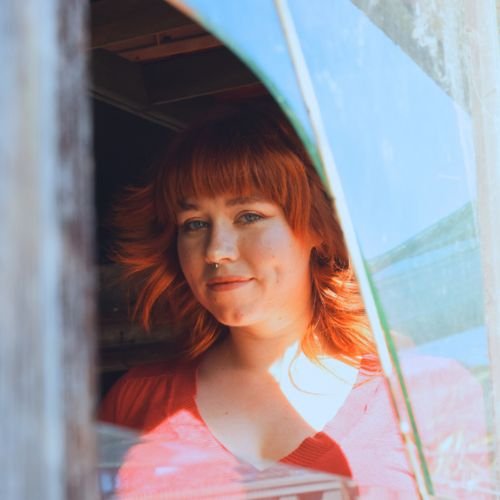 Woman with red hair and bangs holding a colorful umbrella, looking through a window.