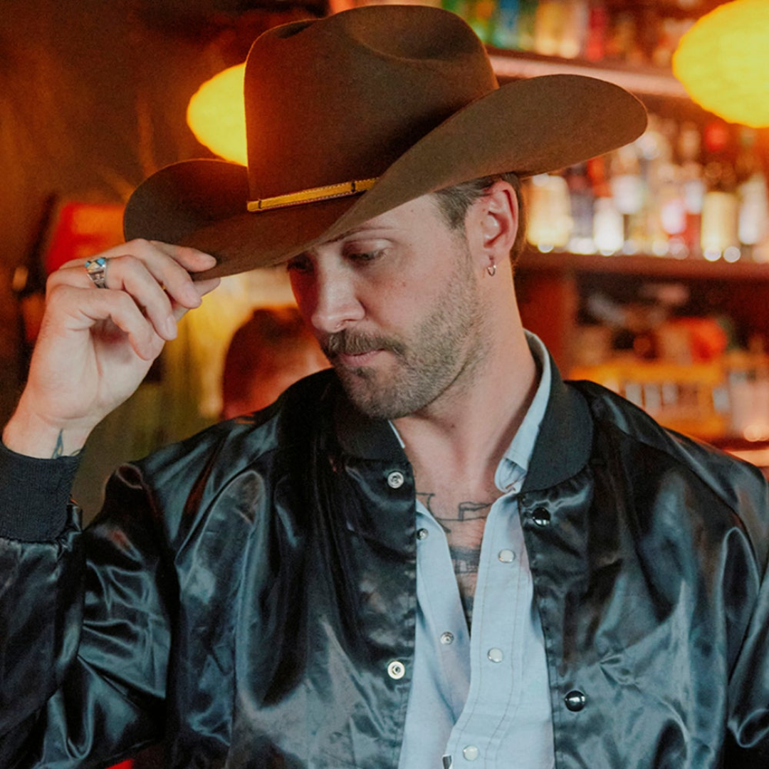 A man wearing a large cowboy hat, a black shiny jacket, and a light-colored shirt, adjusting his hat in a dimly lit bar with bottles and warm lighting in the background.