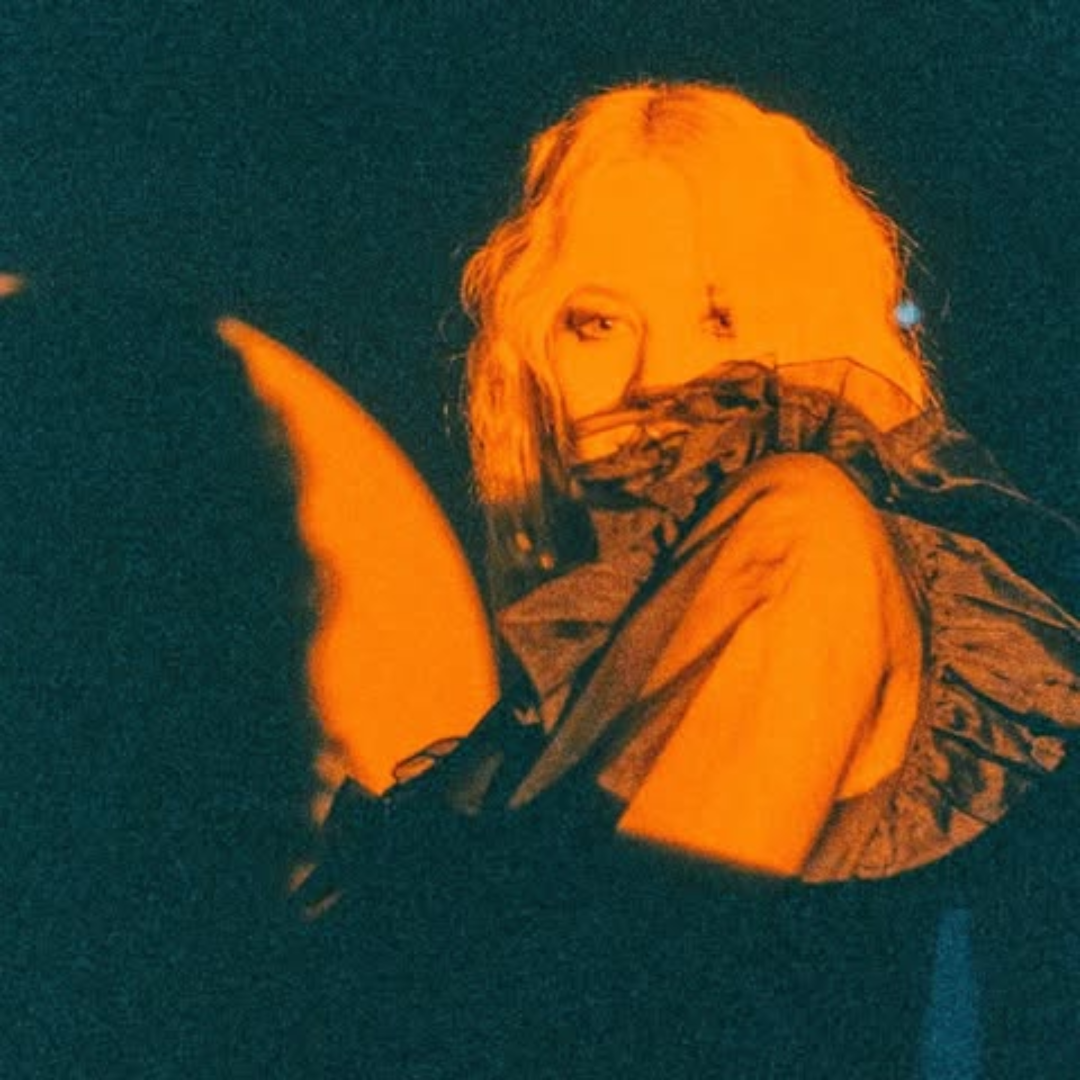 A person with blonde hair and light eyes, wearing a patterned top, looking over their shoulder with one eye visible, in a dimly lit environment with orange and dark tones.