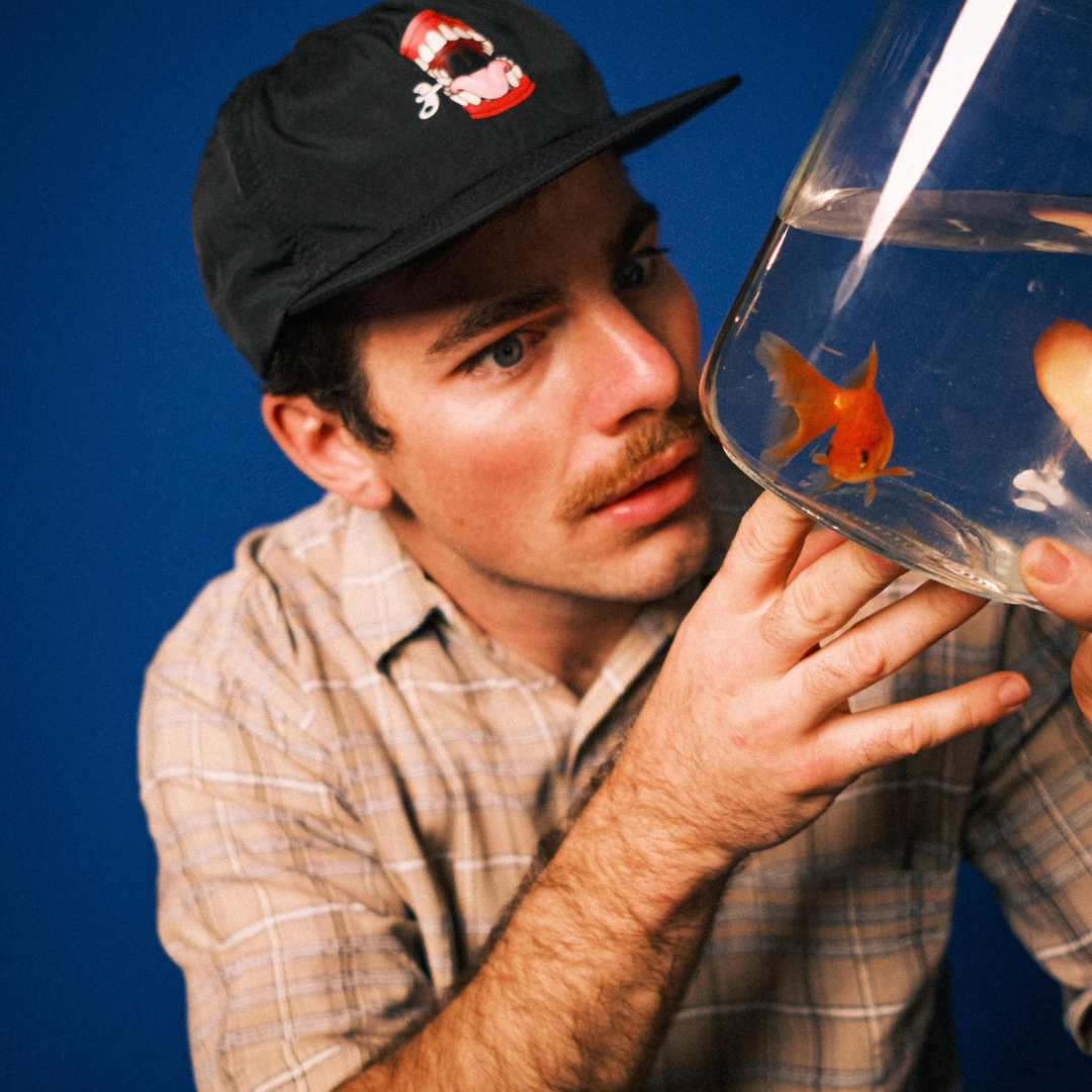 Man with a beige plaid shirt and black cap examining a fishbowl with orange and white fish against a blue background.