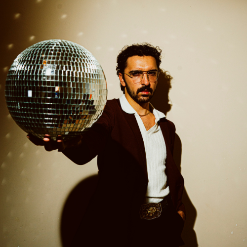 A man with dark curly hair and glasses holding a large disco ball in a room with plain beige walls.