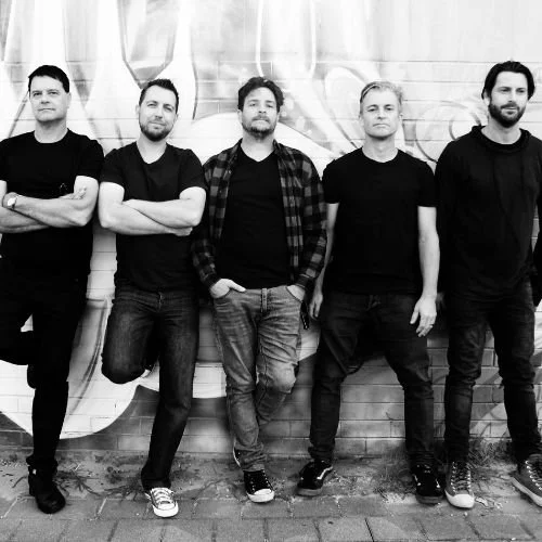 Five men standing in front of a graffiti-covered wall, posing for a photo in black and white.