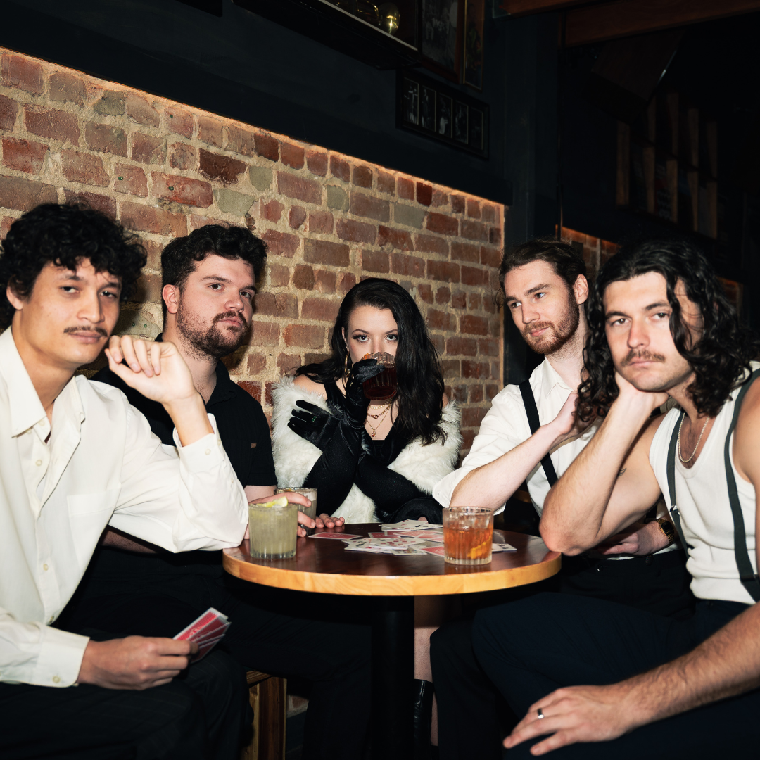 Six young adults sitting around a round table in a dimly lit bar or restaurant with a brick wall background. They are having drinks, some holding glasses, and appear to be posing for the photo.