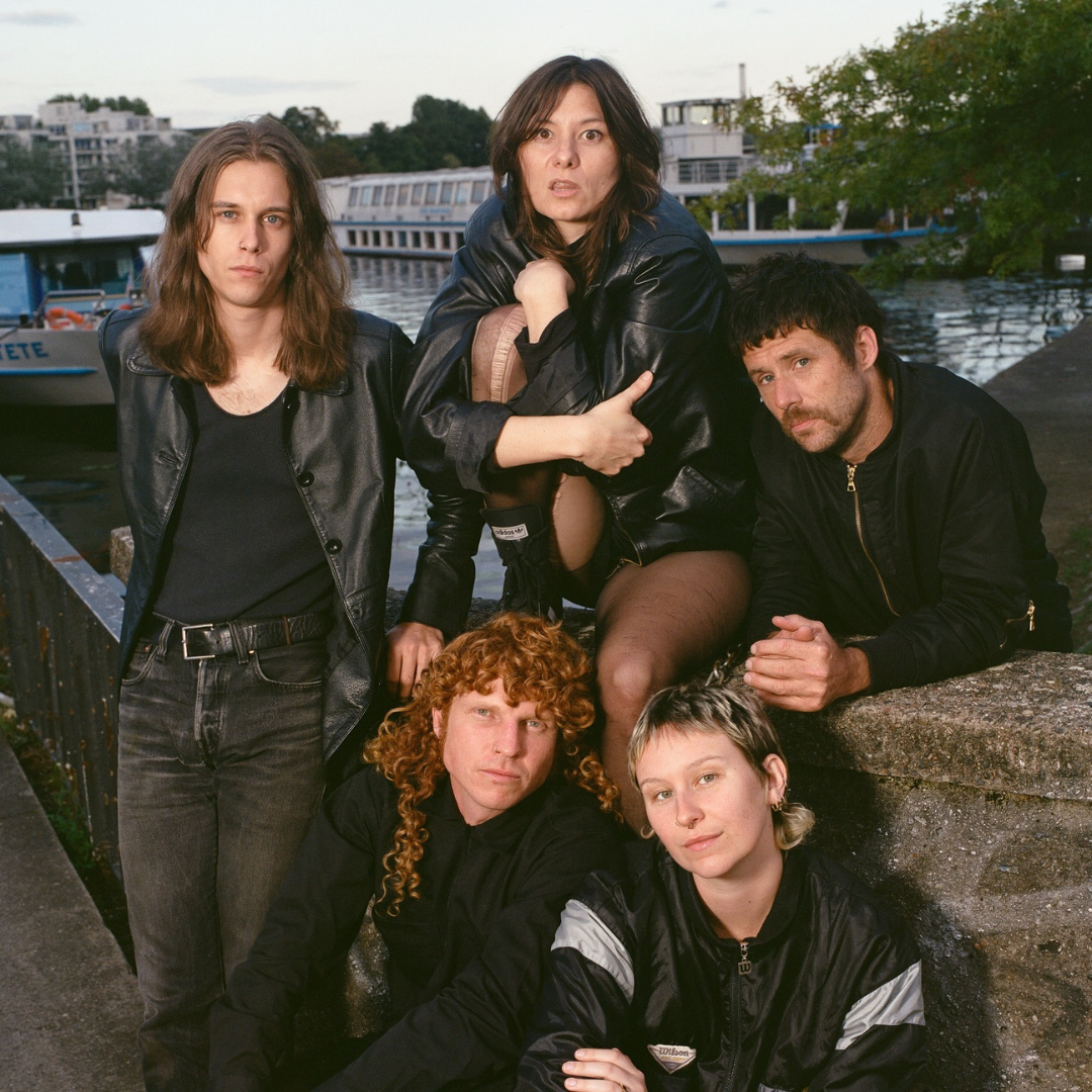 Six young adults, five women and one man, outdoors near a dock with boats and water in the background. They have diverse hairstyles and are dressed in casual black clothing, posing seriously for the photo.