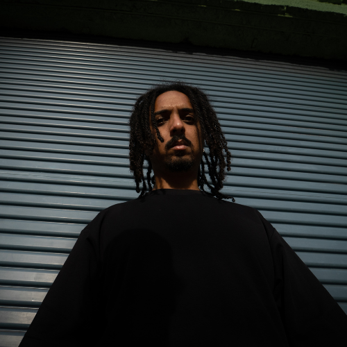 A young man with dreadlocks looking confidently at the camera, standing in front of a metallic roller shutter door.
