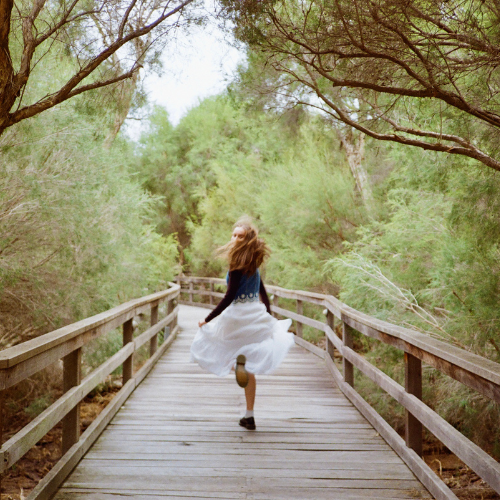 A girl with long hair wearing a white dress running on a wooden boardwalk through a green, wooded area.