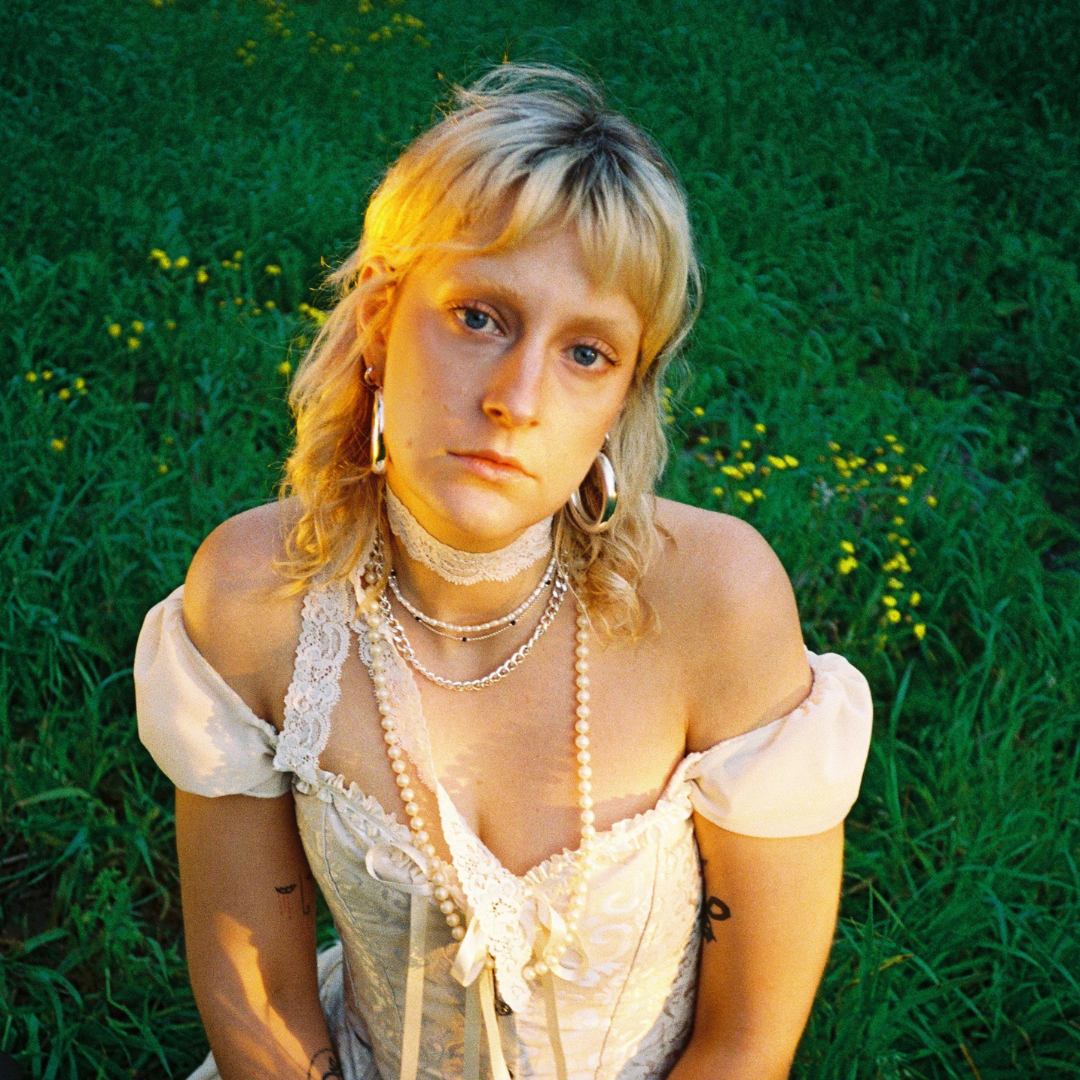 Young woman with blonde hair and blue eyes wearing layered necklaces, earrings, and a lace choker, sitting in a grassy field with yellow flowers.