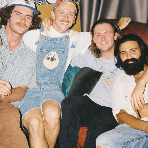 Four men sitting close on a sofa, smiling, with their arms around each other. They appear to be friends in a casual setting.