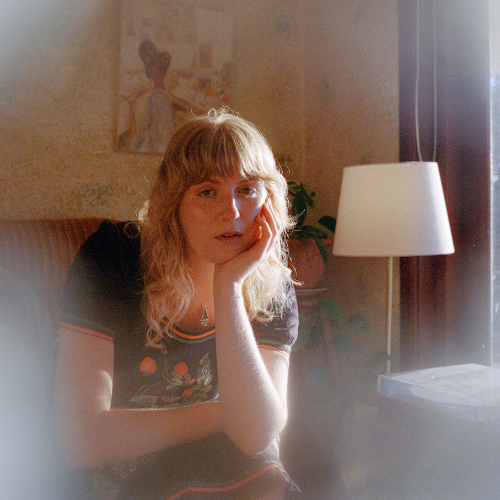 A woman with blonde hair sitting at a table, resting her head on her hand, next to a table lamp, with a window and a painting in the background.