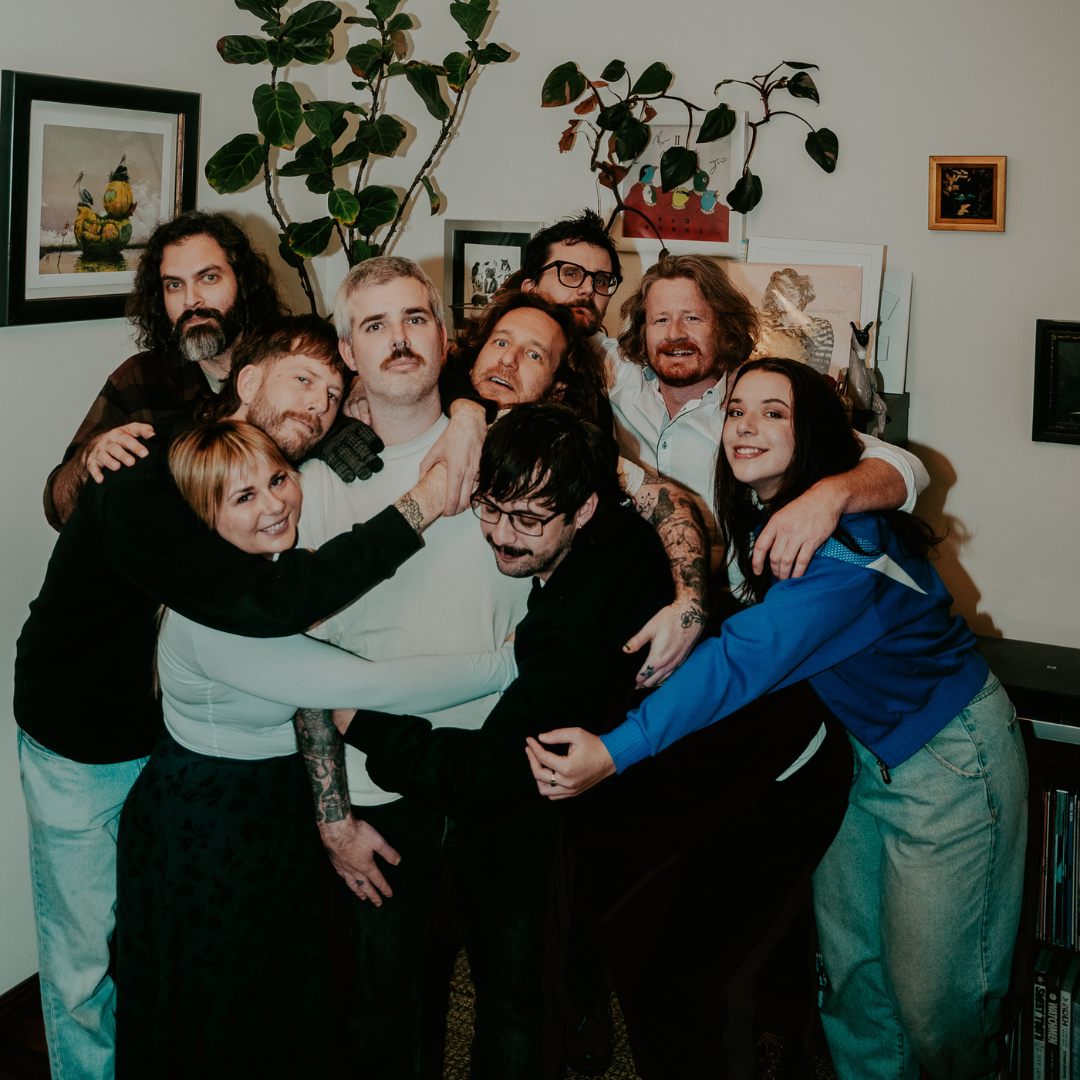 Group of nine diverse people hugging and smiling together in a living room, with art and plants in the background.
