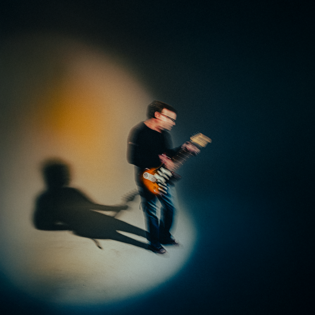 Blurred image of a person playing an electric guitar, with their shadow cast on the wall behind them.