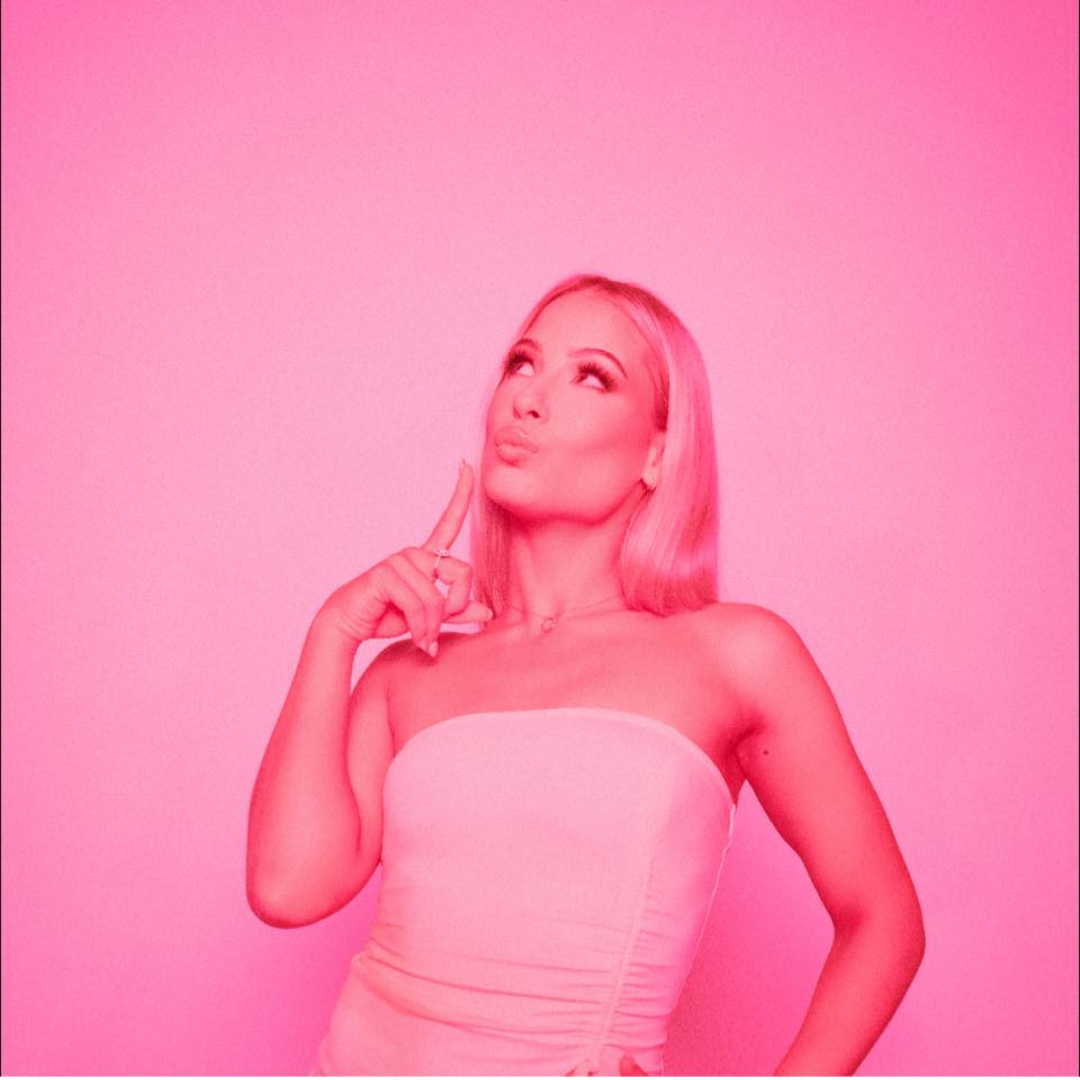 A woman with blonde hair in a strapless dress, posing against a pink background with her finger on her chin, looking upwards thoughtfully.