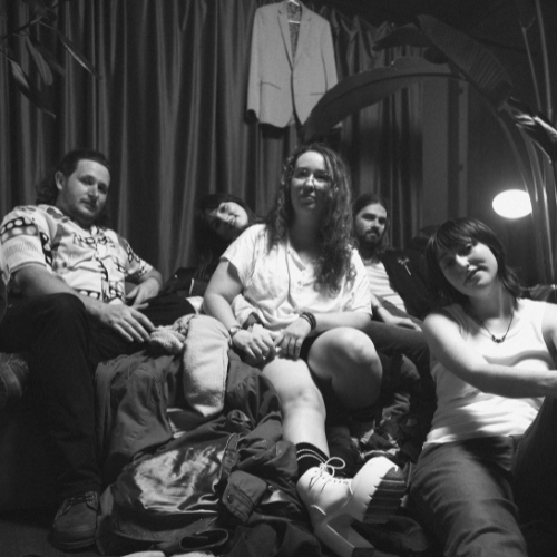 Group of four young adults sitting on a couch in a room with a curtain backdrop, with a coat hanging on the curtain and a large plant behind them. The scene is in black and white.