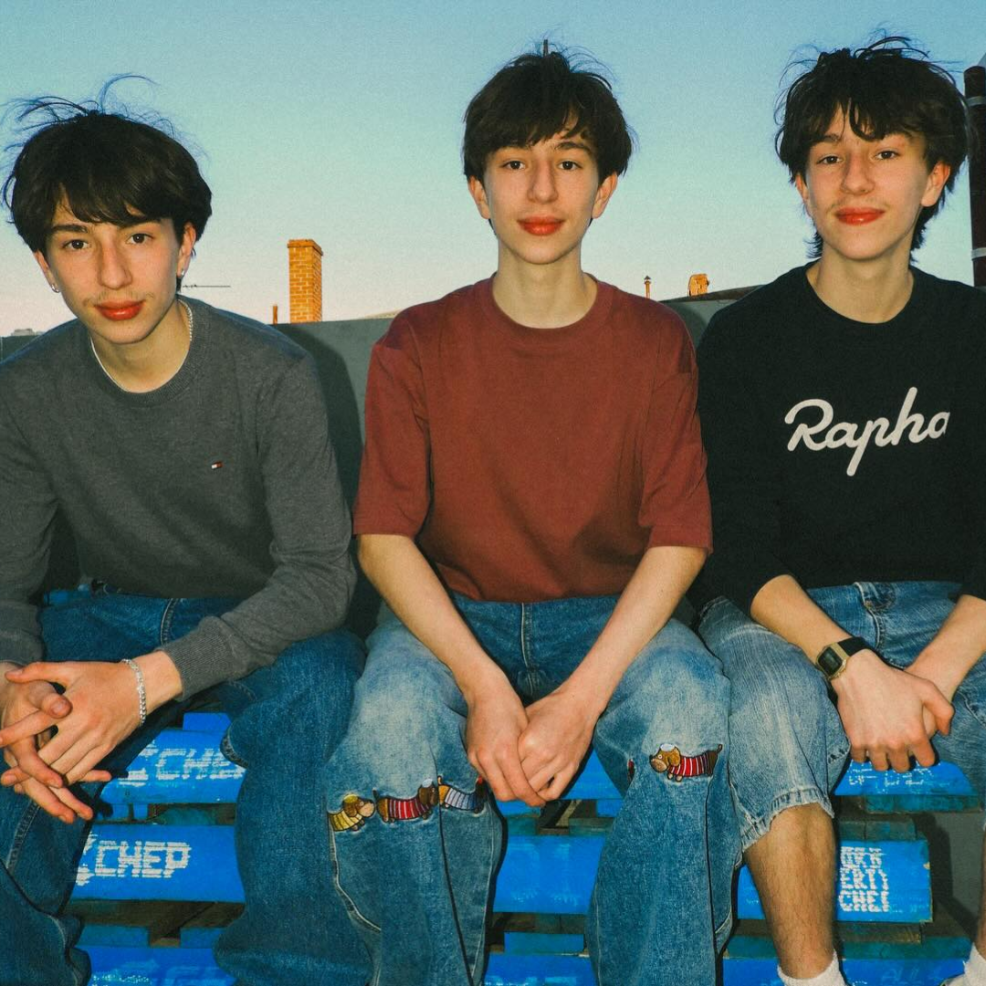 Three young men with dark hair sitting on a bench outdoors during the day, smiling at the camera.