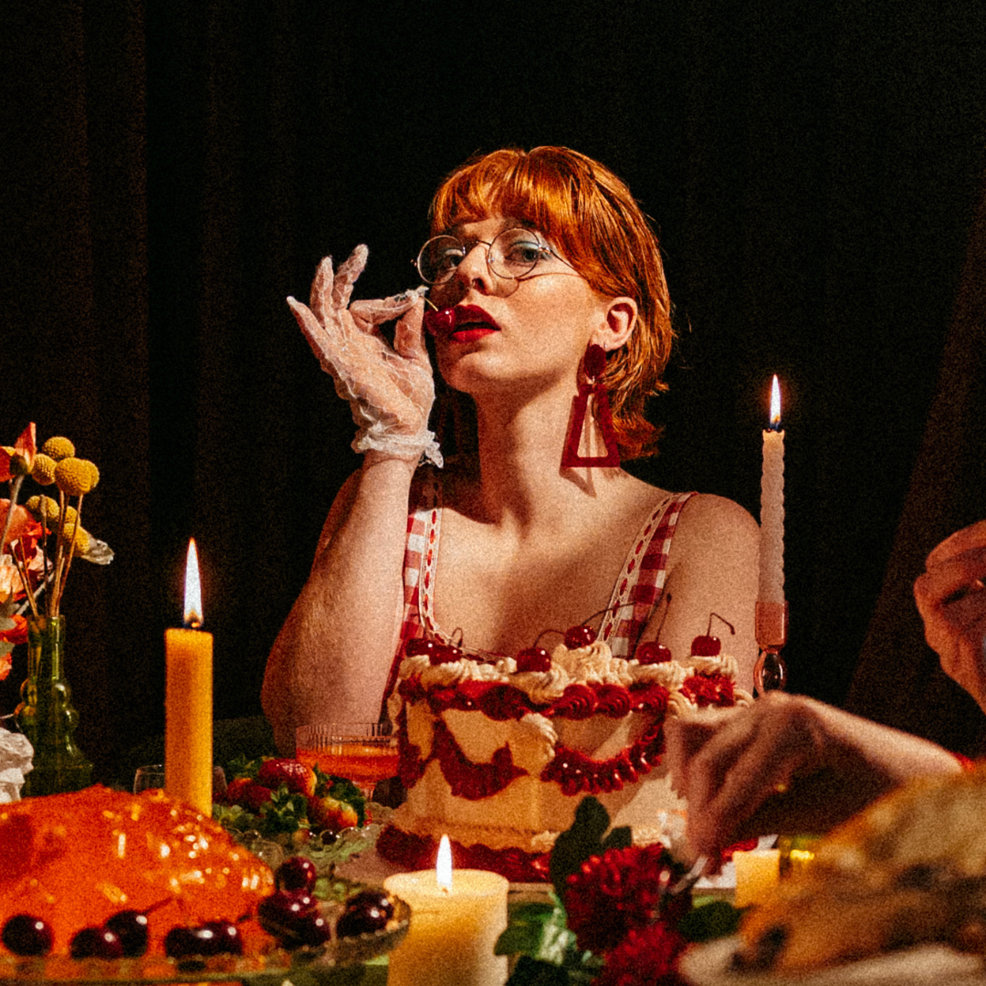 A woman with red hair and glasses sitting at a table with cakes and candles, wearing a red and white checkered dress and large red earrings, in a dimly lit setting.