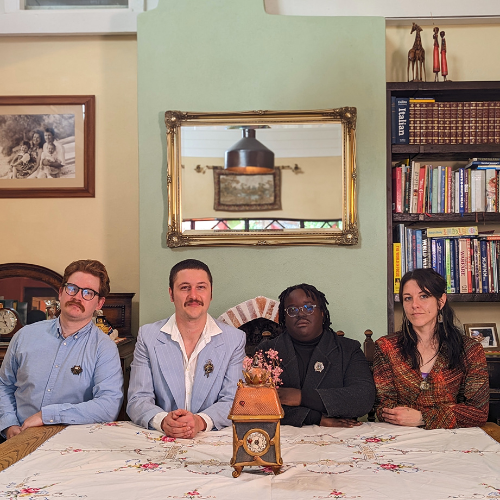 Four people sitting at a dining table in a room with bookshelves, mirror, and framed picture on the wall.