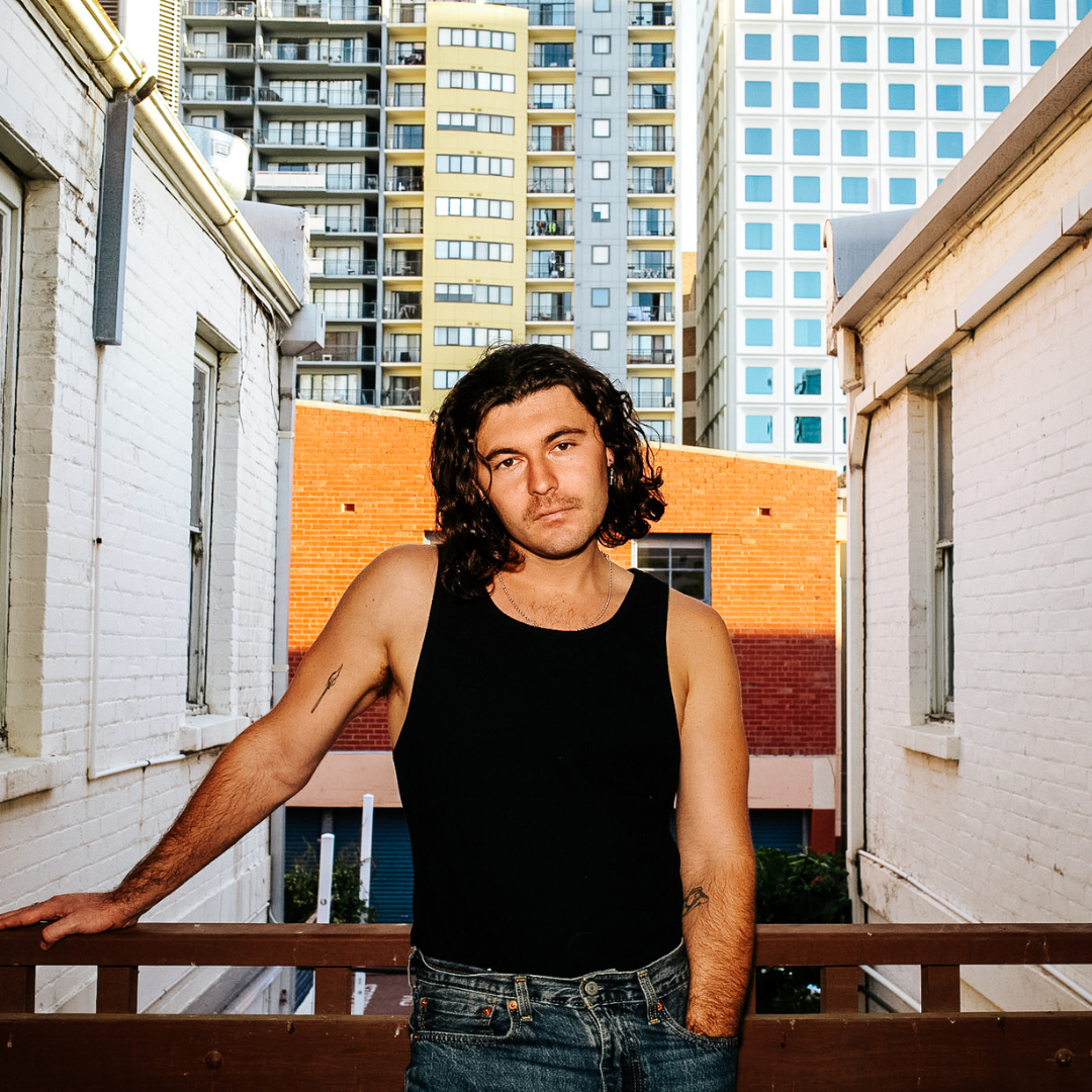 A young man with curly dark hair, wearing a black tank top and jeans, standing outdoors on a balcony with urban high-rise buildings in the background.