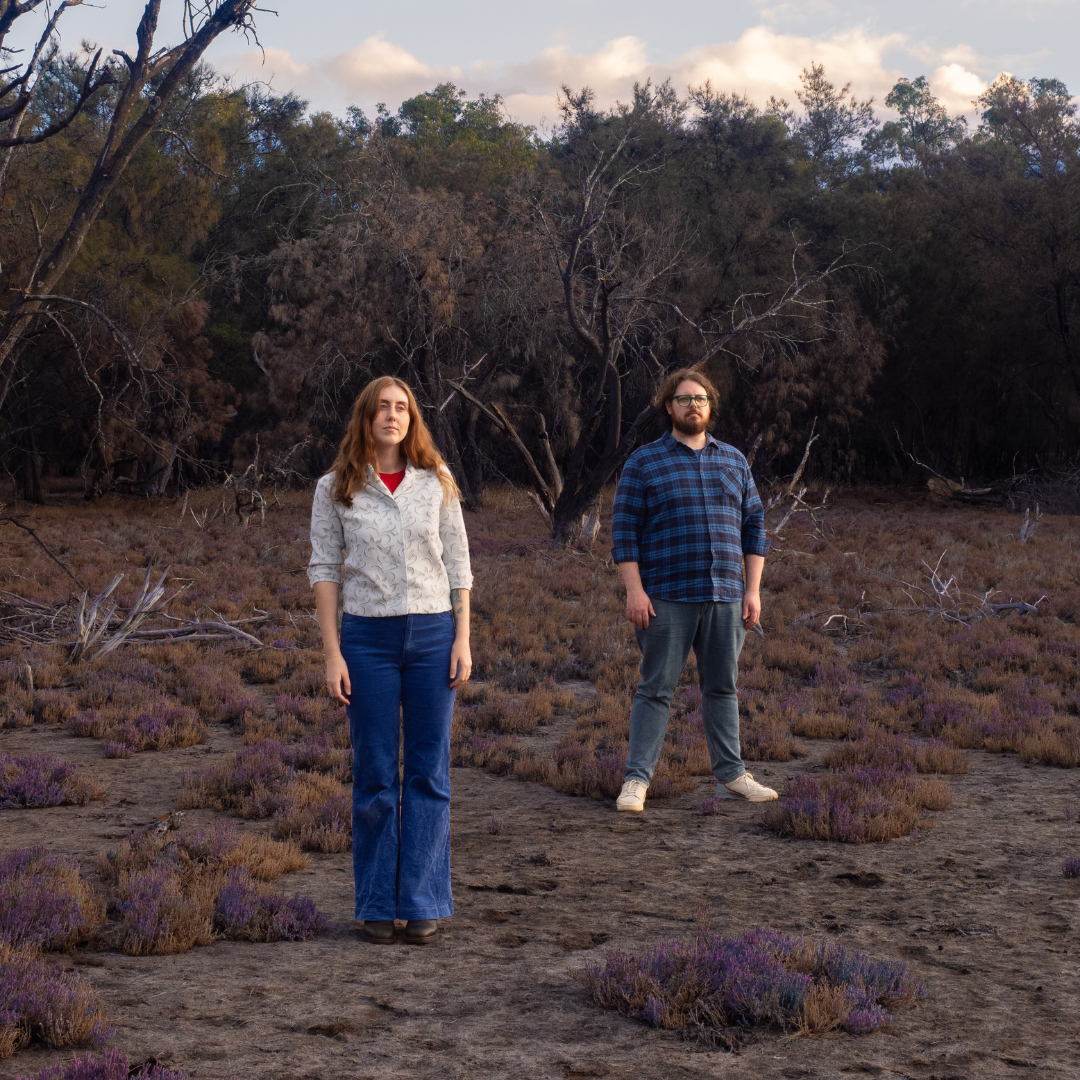 Two people standing in a field with purple plants and bare trees in the background, under a cloudy sky.