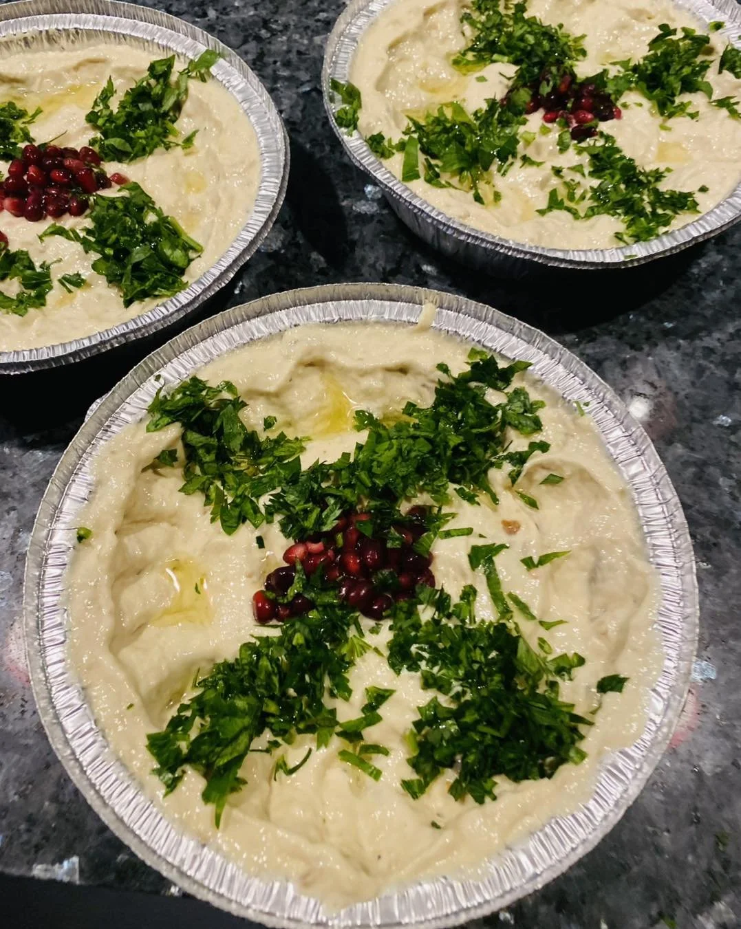 Three aluminum foil containers filled with a creamy dip garnished with chopped parsley, chopped green onions, and pomegranate seeds, placed on a dark granite countertop.