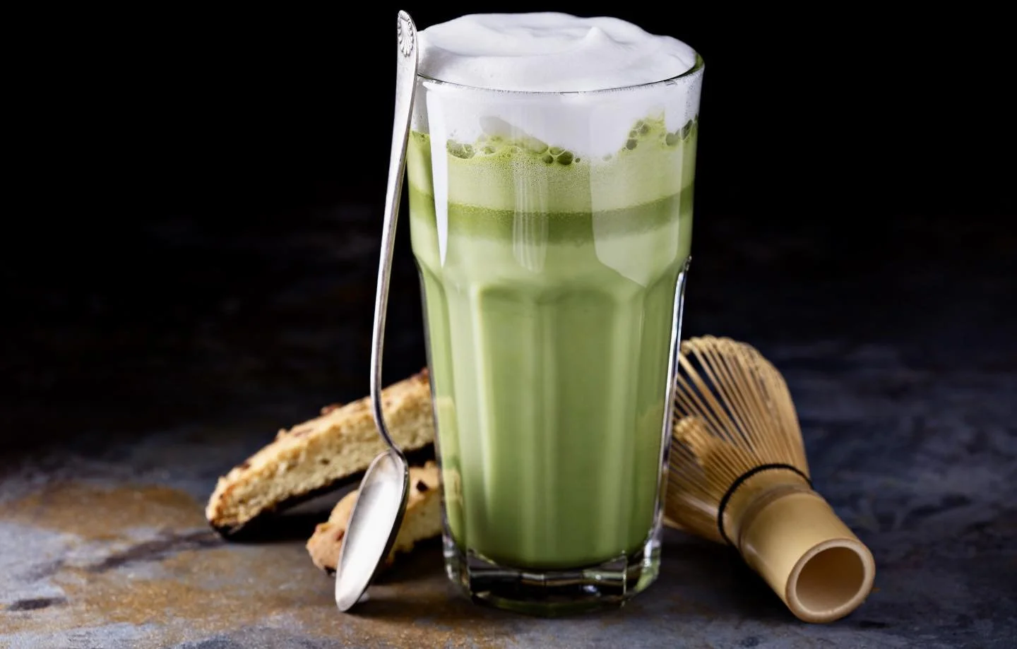 A tall glass of green matcha latte with whipped cream on top, accompanied by biscotti cookies, a bamboo matcha whisk, and a silver spoon, set on a dark surface.