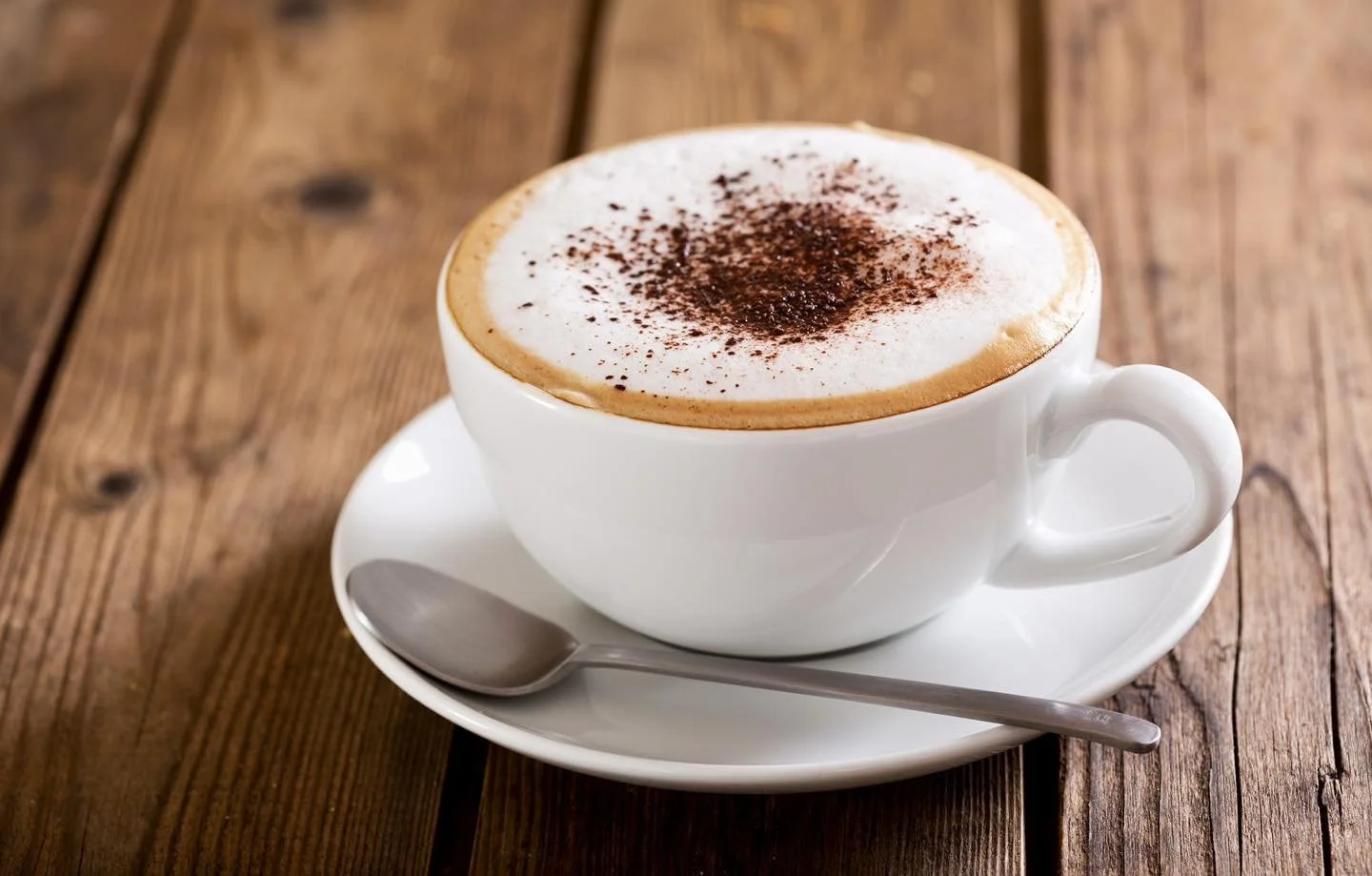 A cup of cappuccino with foam and cocoa powder on top, placed on a white saucer with a spoon, on a wooden table.