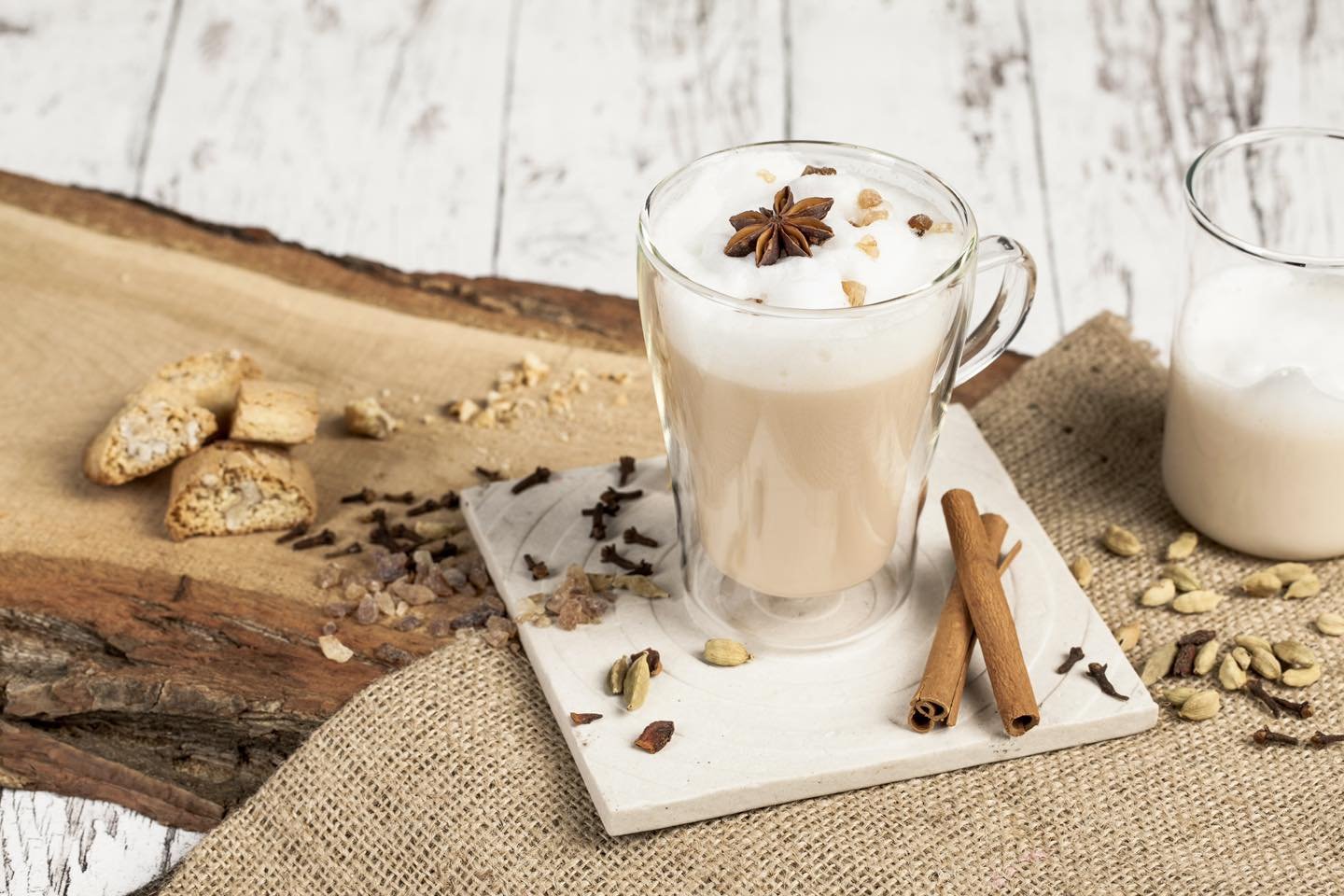 A glass mug filled with a creamy coffee beverage topped with whipped cream, star anise, and crushed nuts. Surrounding the mug are cinnamon sticks, cardamom pods, and scattered spices on a rustic wooden surface and burlap cloth.