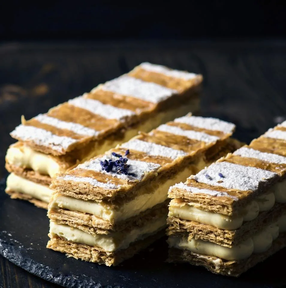 Layered pastry dessert with white icing, powdered sugar, and a small cluster of purple edible flowers on top, served on a black slate platter.