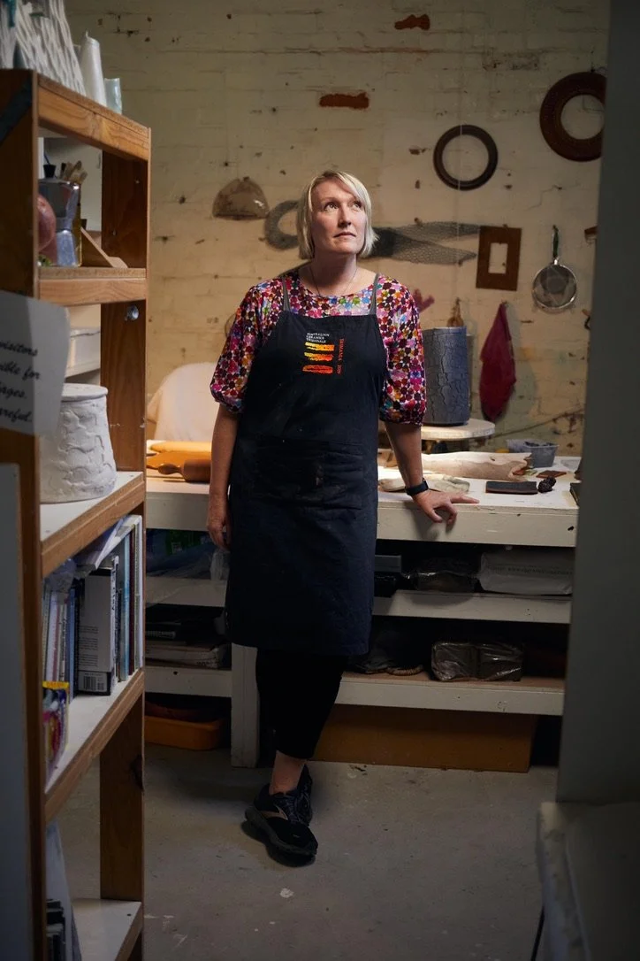 A woman with blonde hair wearing a colorful patterned shirt, black apron, and black sneakers stands in a pottery studio. She leans against a worktable with pottery tools and objects, with a rustic wall behind her decorated with frames and hanging tools.