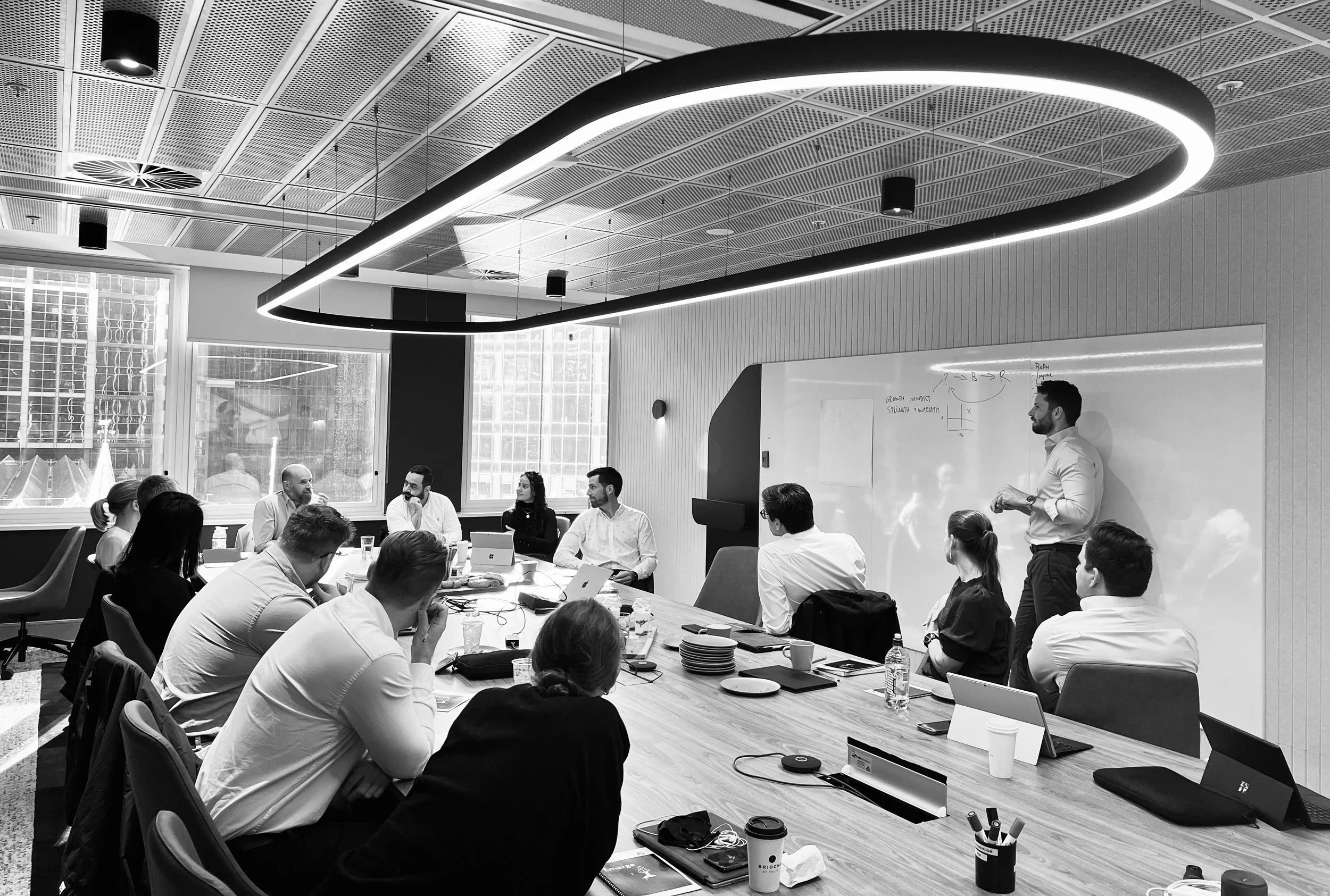 A group of people in a modern conference room participating in a meeting, with a man giving a presentation at a whiteboard.
