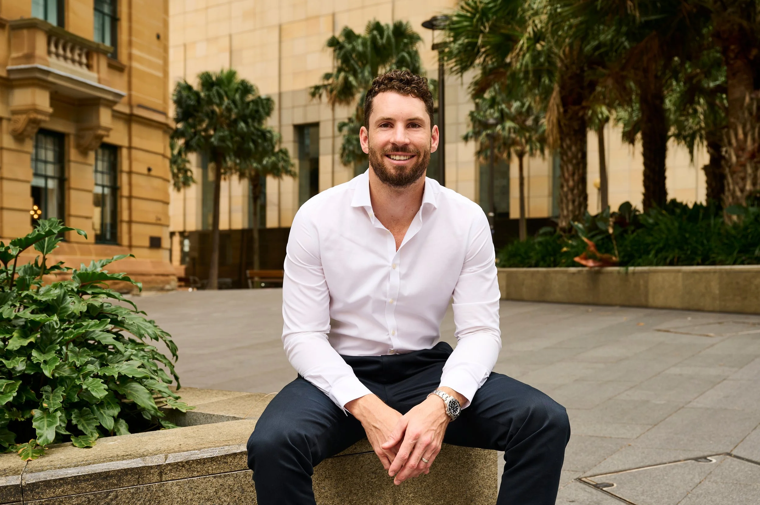 A smiling man in a white dress shirt and dark pants sitting on a stone bench outside in an urban setting with palm trees and a historic building in the background.