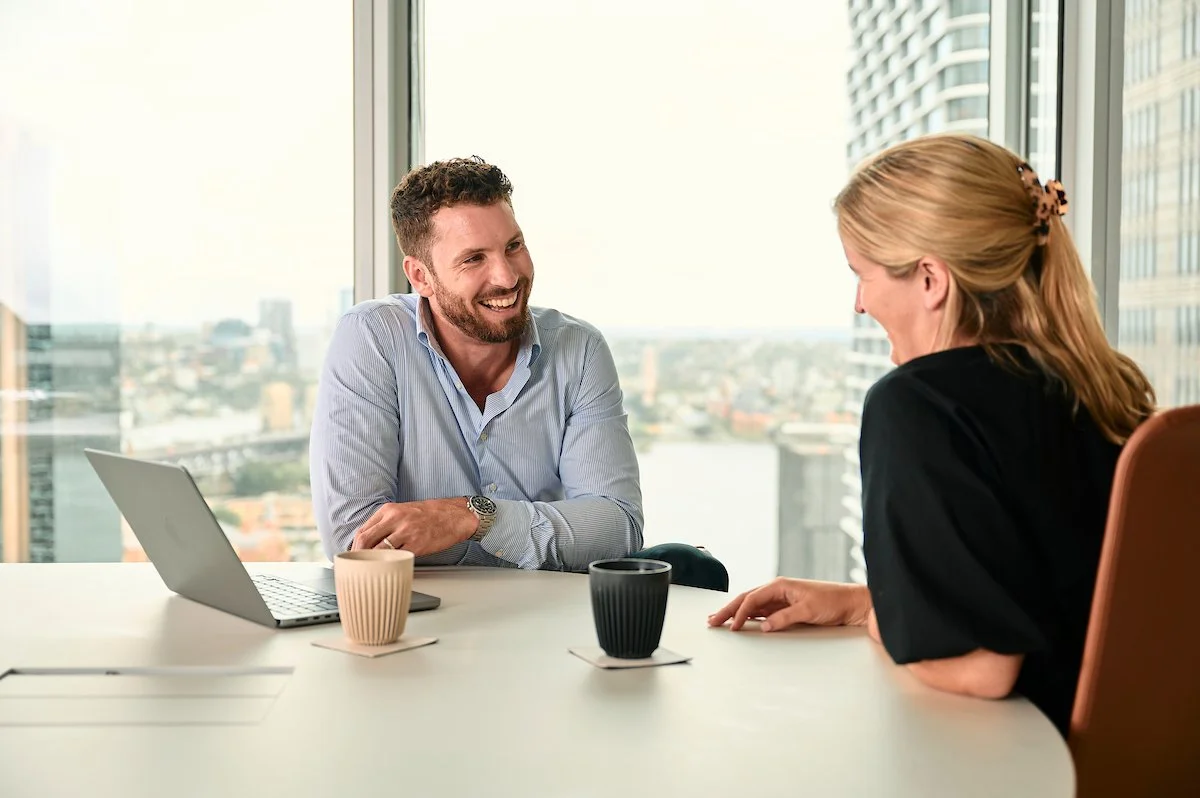 A man and woman having a conversation in a modern office with a view of the city through large windows.