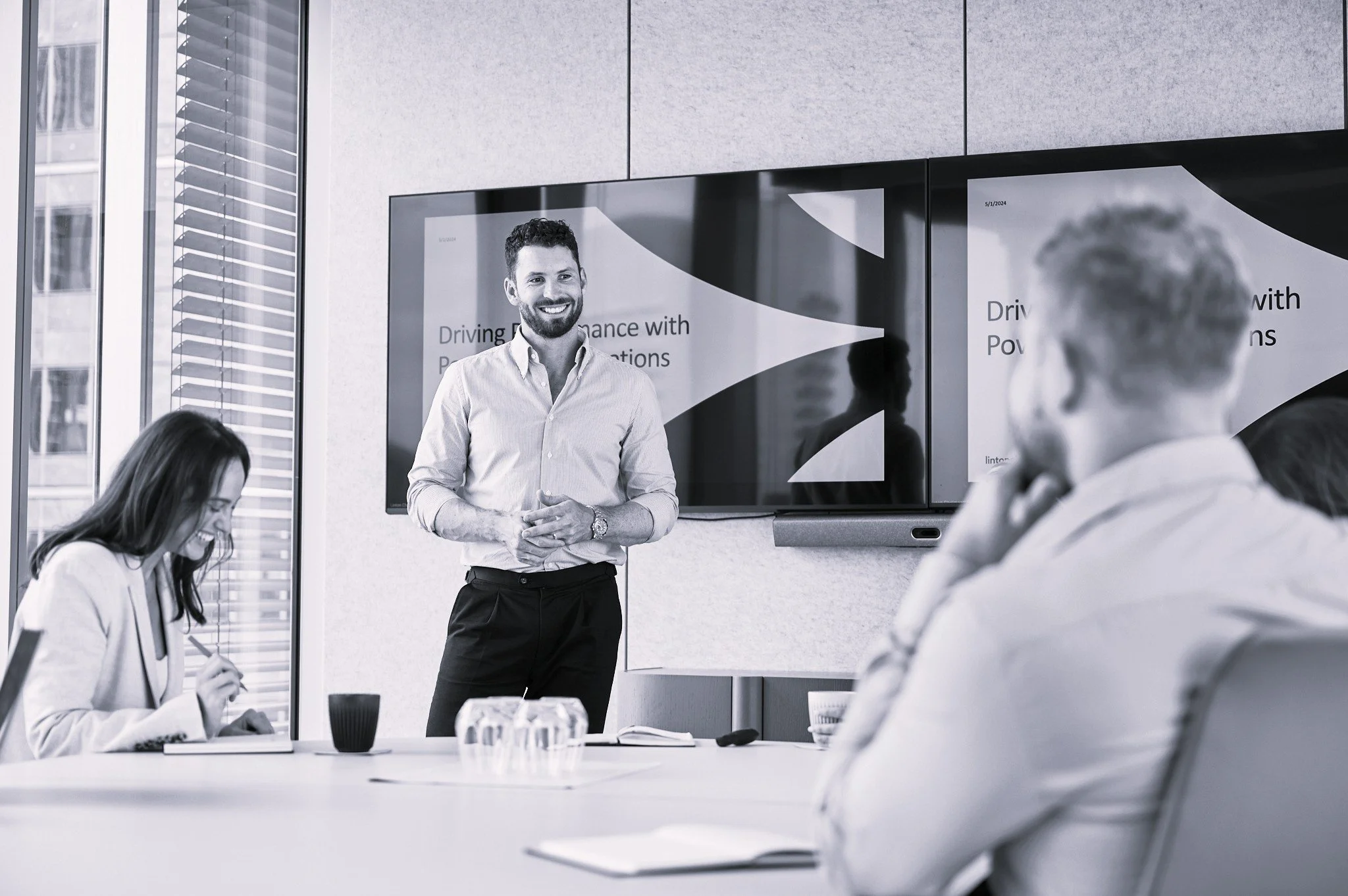 Business meeting in office with man giving presentation and others listening, large screen displays presentation slides.