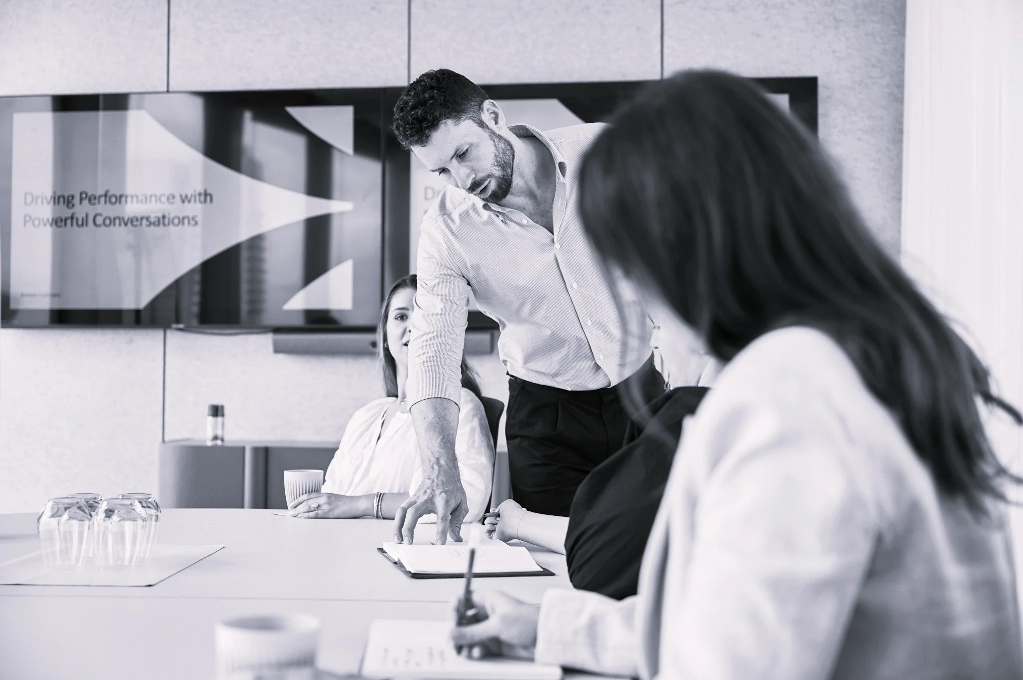 A man standing and leaning over a conference table, engaging in a discussion with three women seated at the table in a modern conference room. There are three glasses of water and notebooks on the table, with a large screen displaying text in the background.