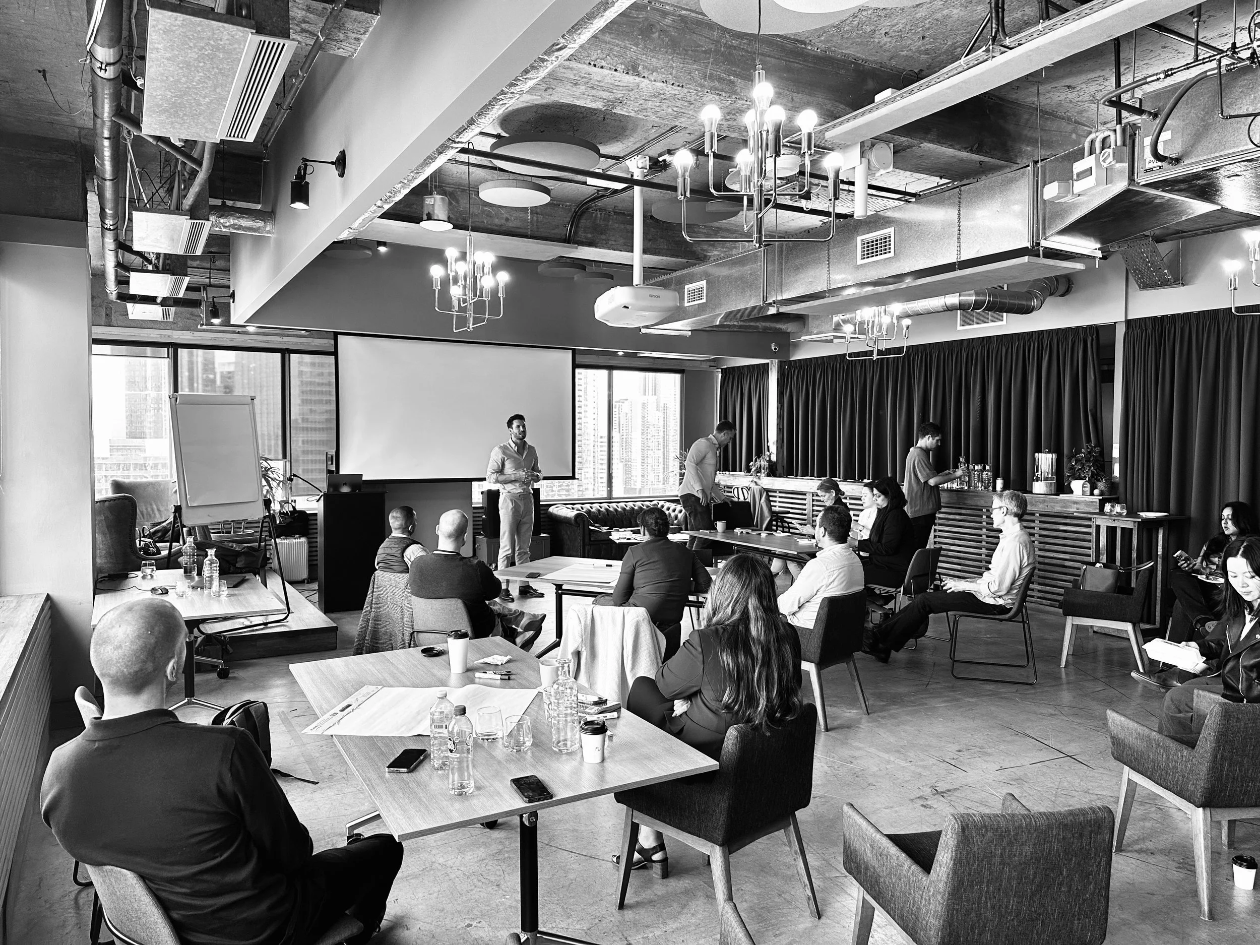 A business presentation or meeting with a speaker at the front, seated attendees, and a bar area with drinks; black and white photo.