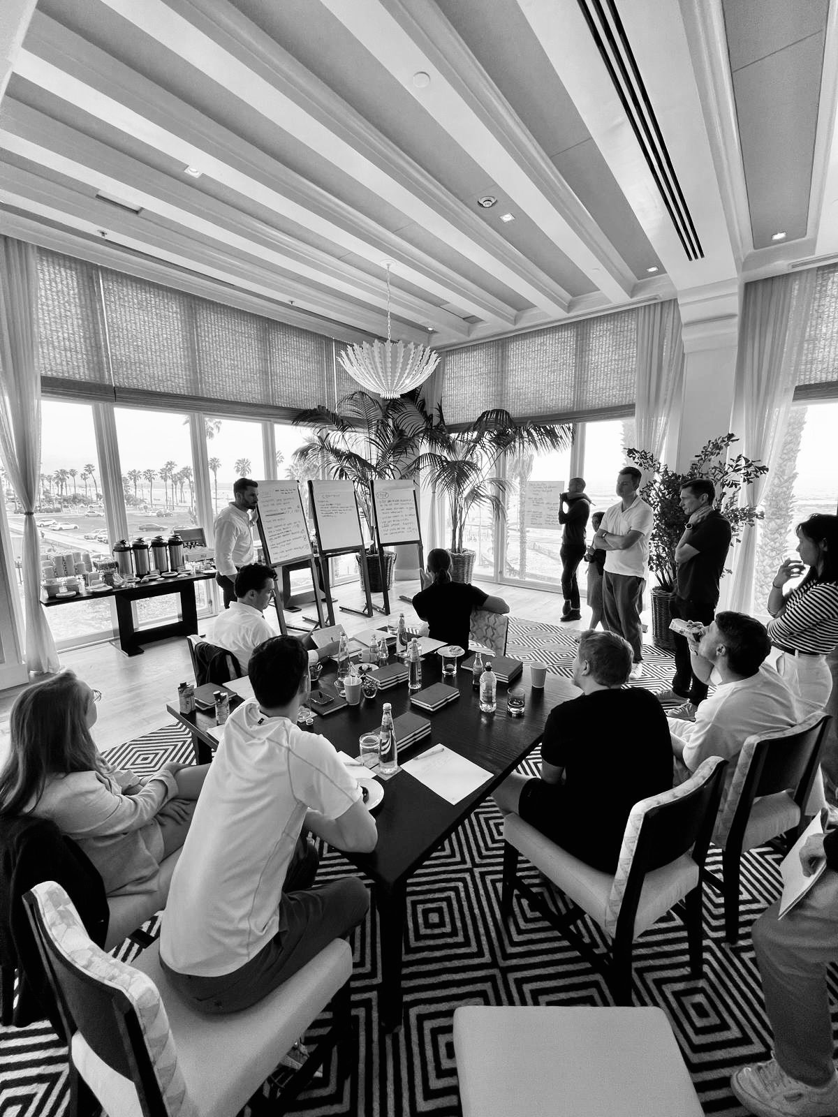 A black and white photo of a group of people in a conference room with large windows, tables, chairs, and presentation boards, during a meeting or workshop.