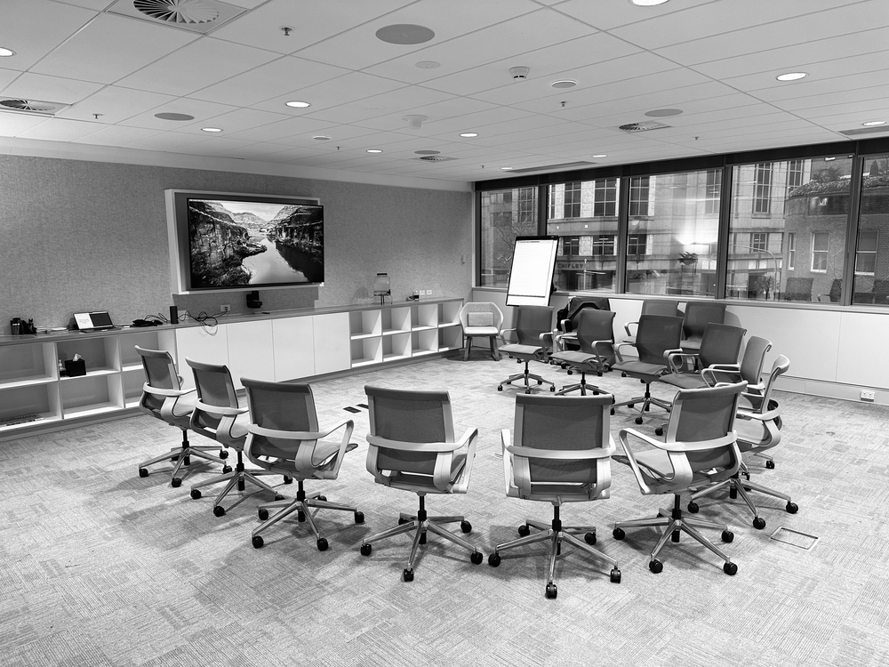 Empty conference room with eleven chairs arranged in a semi-circle, a large wall-mounted TV displaying a landscape, a whiteboard on an easel, and large windows showing city buildings outside.