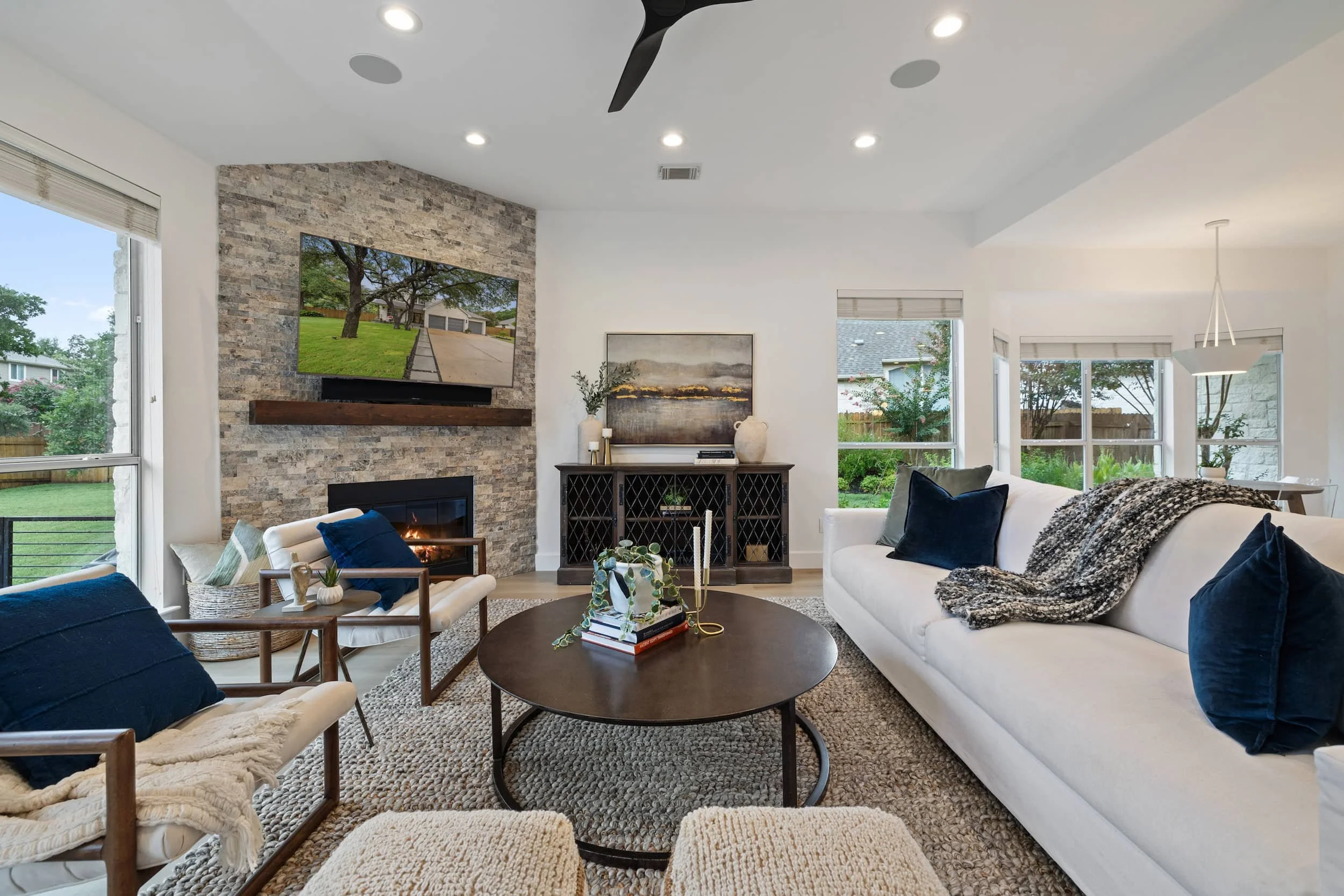 Open living room with stone fireplace and TV, beige sofa and chairs, round coffee table, and large windows with backyard views.