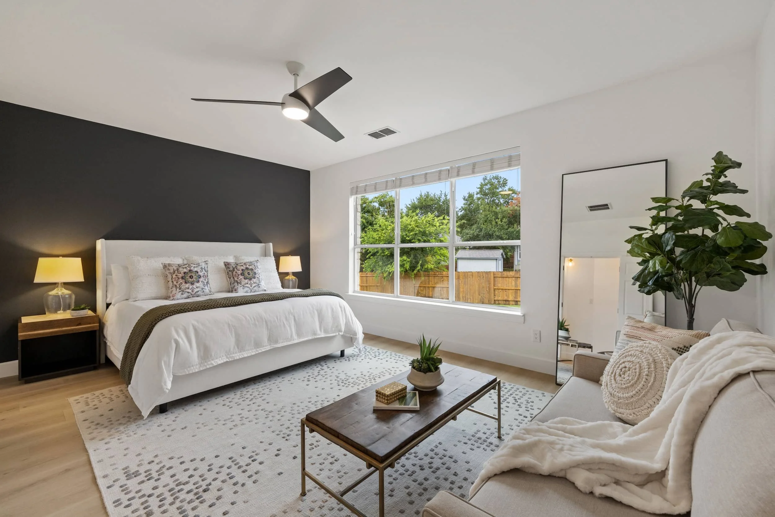 Bedroom with dark accent wall, white upholstered bed, large window overlooking trees, wooden nightstand, full-length mirror, and light gray rug.