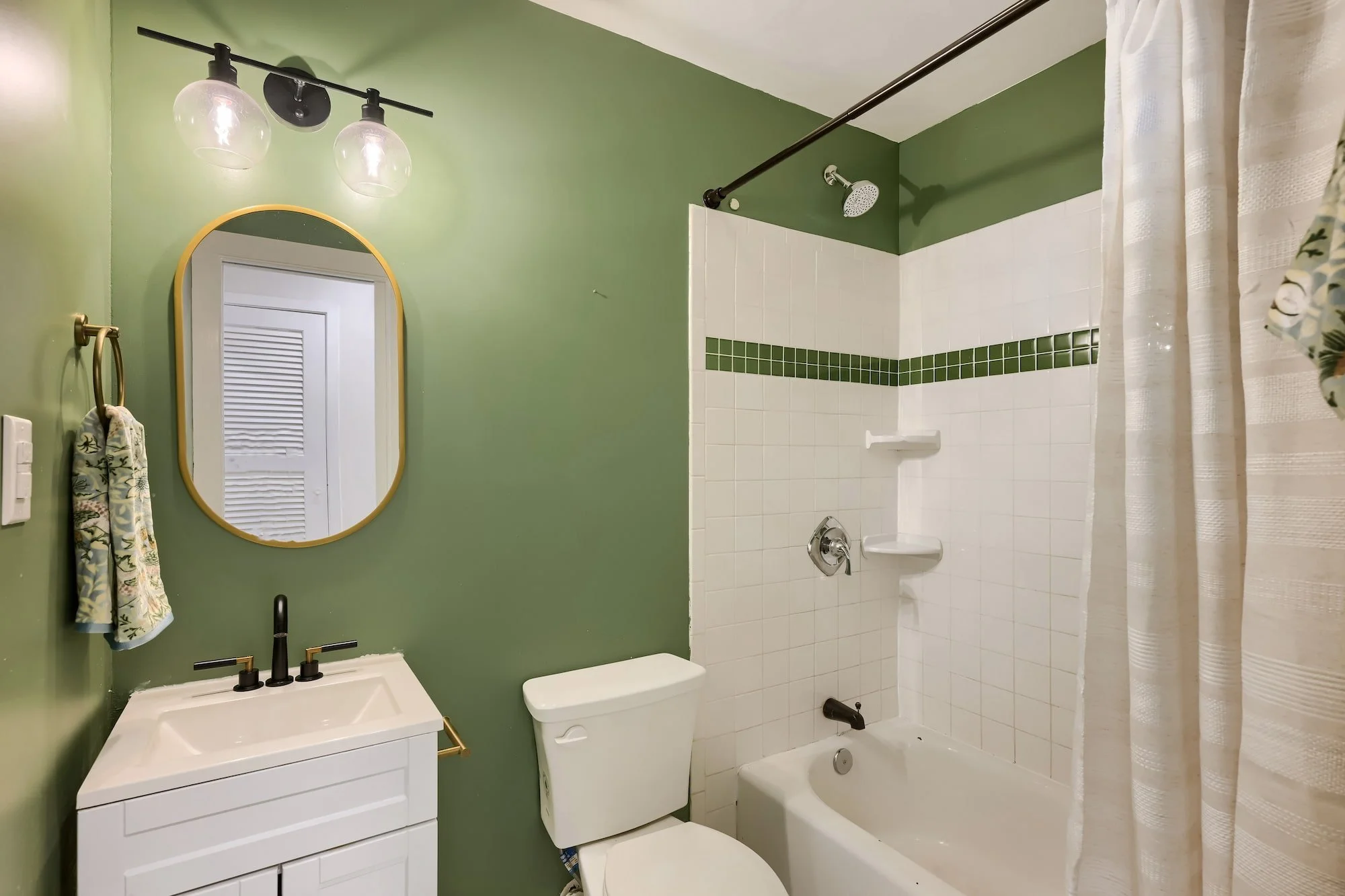 Staged bathroom with sage green walls, a white tub and shower with green tile accent stripe, a white vanity, and a gold oval mirror.