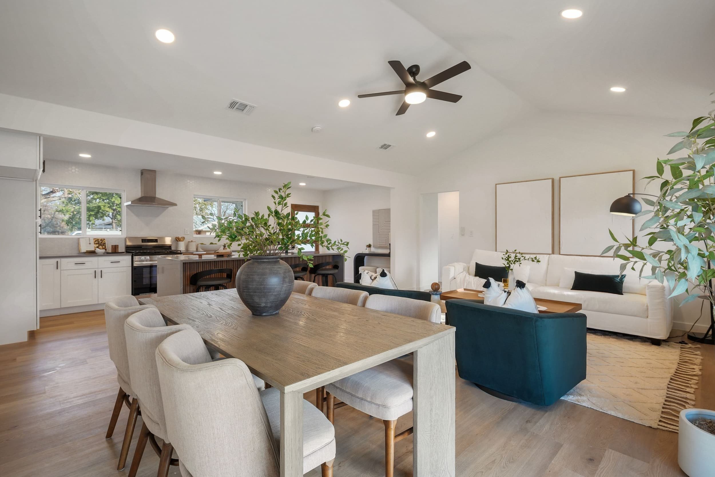 Open-concept living and dining area with wood table, kitchen island, white sofa, teal chairs, ceiling fan, and natural light.