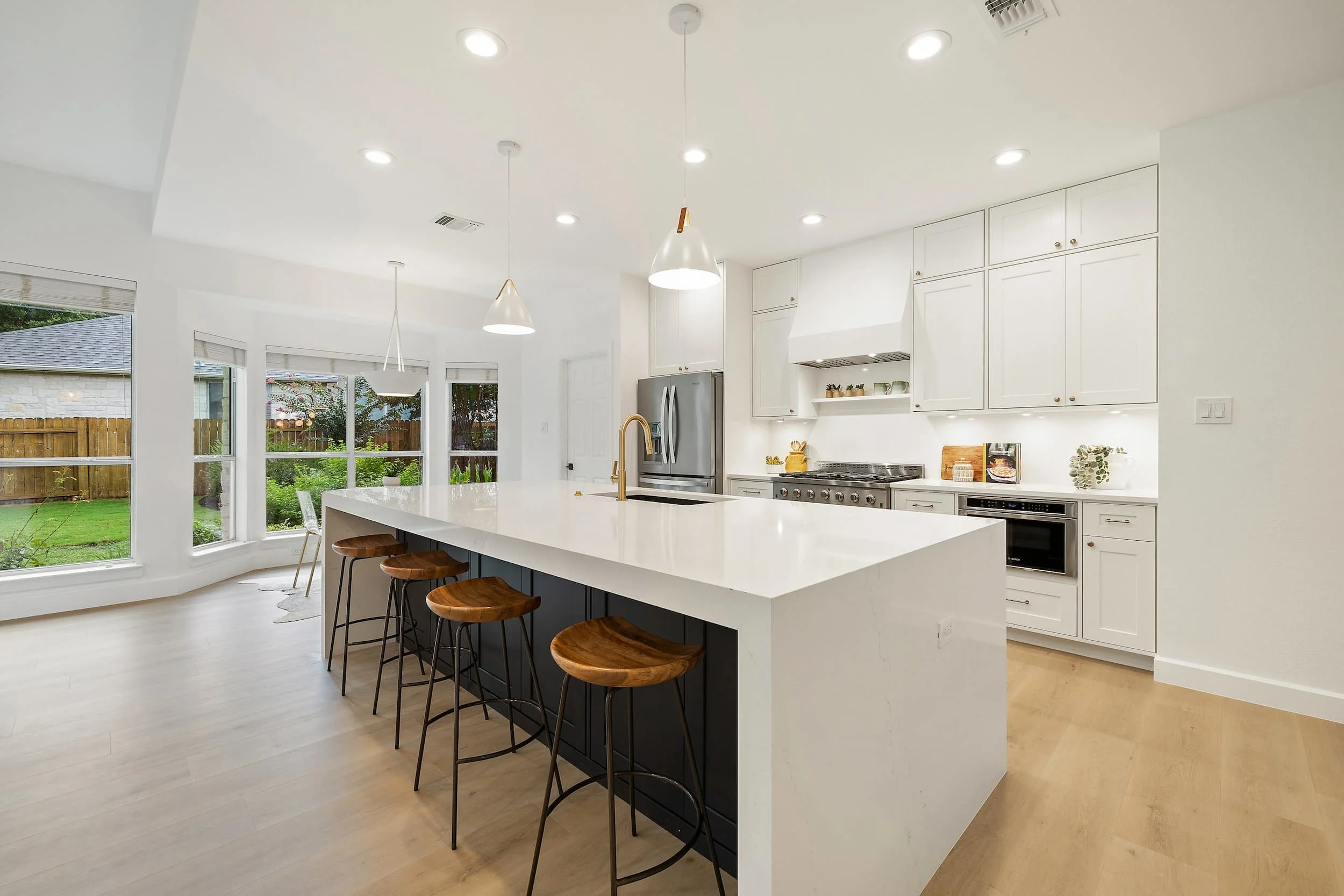 Spacious modern kitchen with a large white island, wooden bar stools, pendant lighting, and white cabinets, with windows overlooking a backyard.