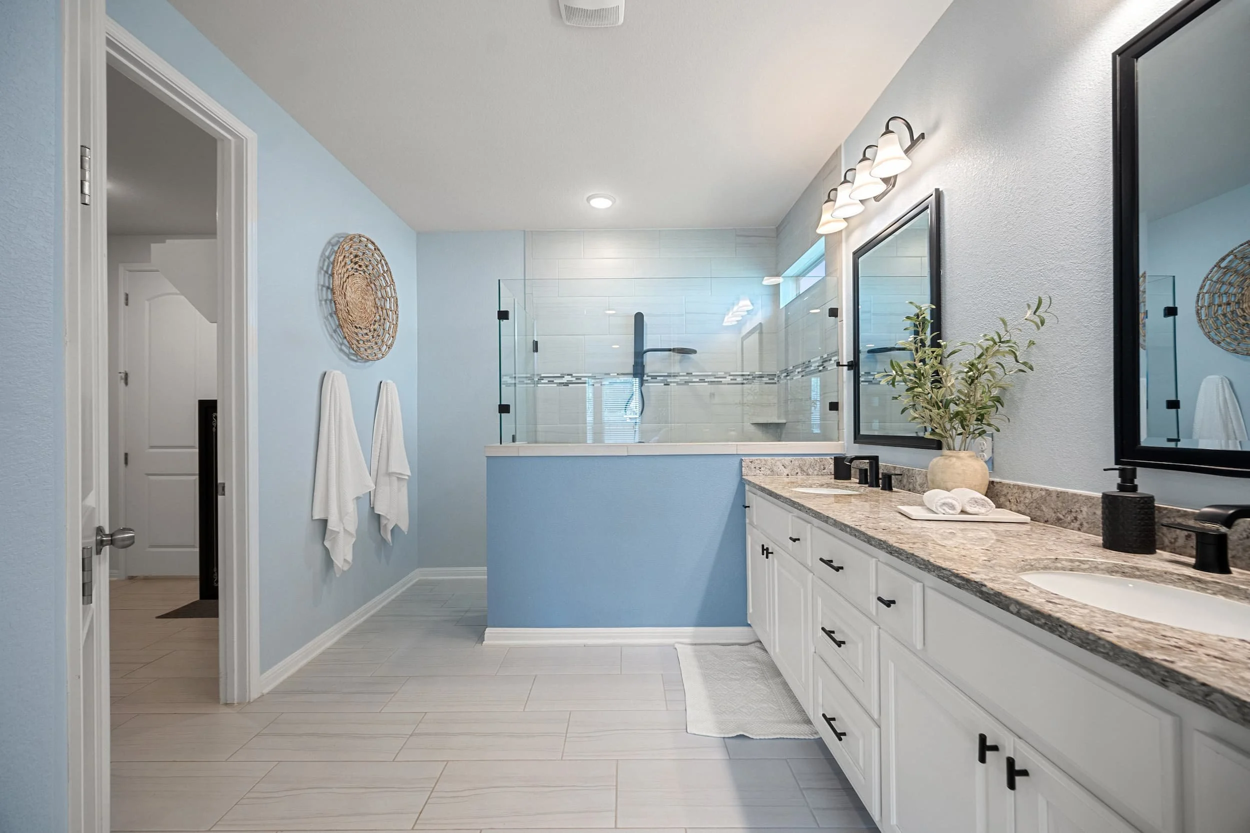 Staged bathroom with light blue walls, white double vanity, granite counters, and a glass-enclosed shower.
