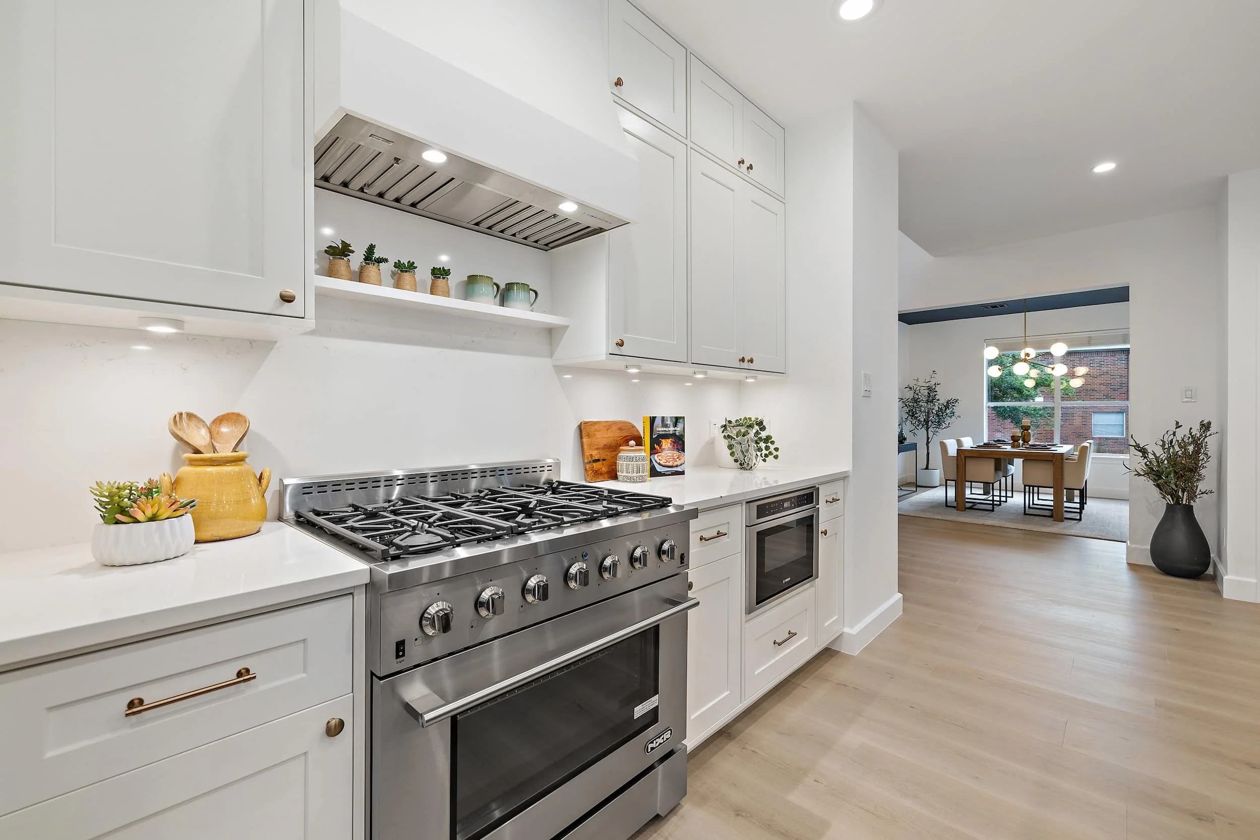 Modern kitchen with white cabinets, a stainless steel gas range and oven, under-cabinet lighting, and light wood floors, opening toward a dining area.