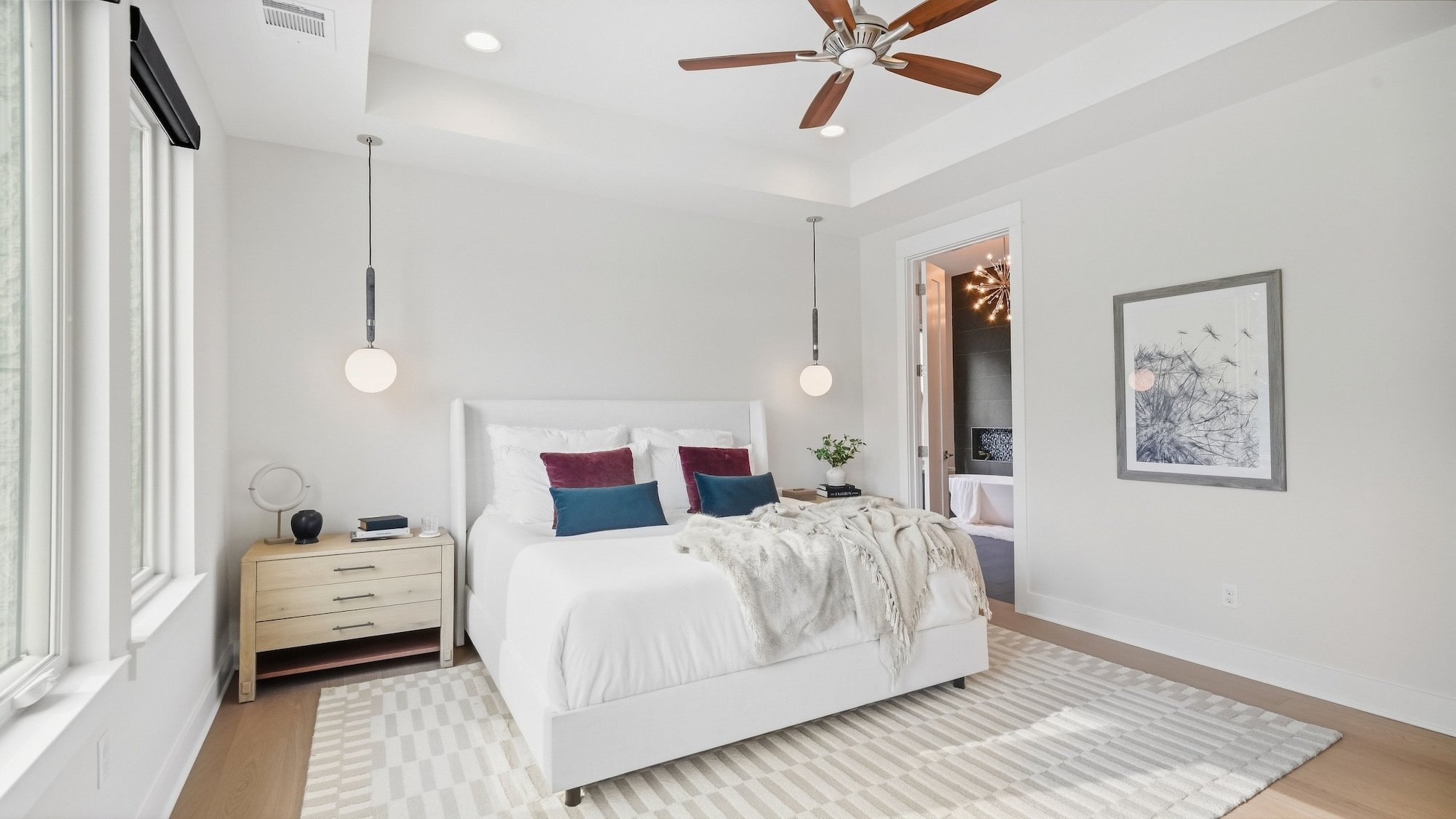 Primary bedroom staged with a white upholstered bed and dandelion art, opening to the primary bathroom.