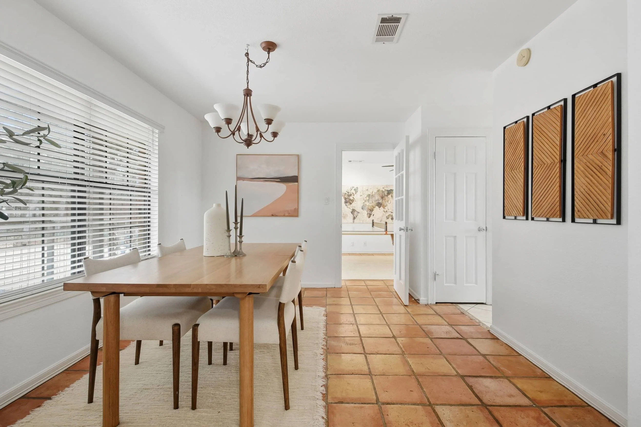 Bright dining room with wood table, upholstered chairs, chandelier, large window, and tile flooring.
