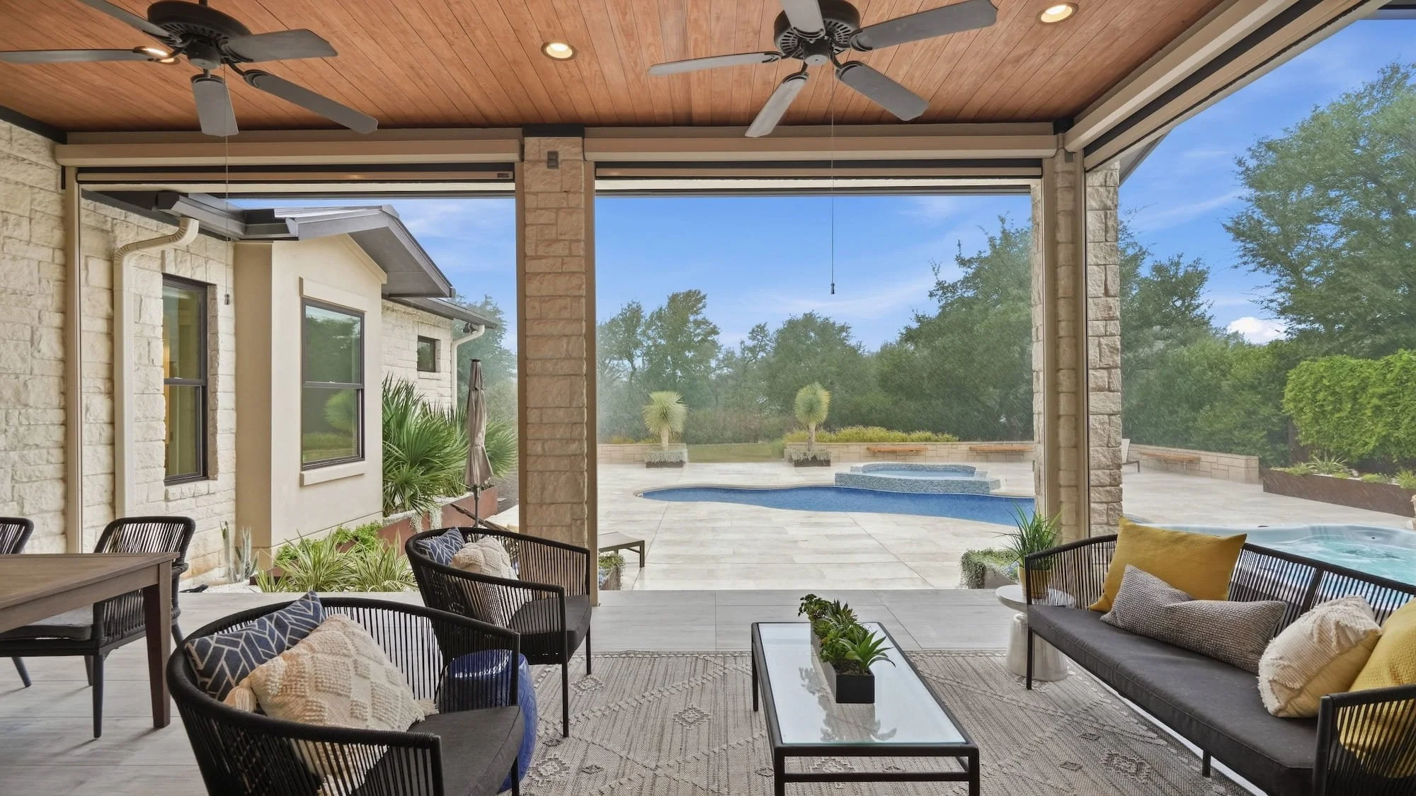 Covered patio staged with black rope chairs, a sofa with yellow pillows, and a glass coffee table overlooking the pool.